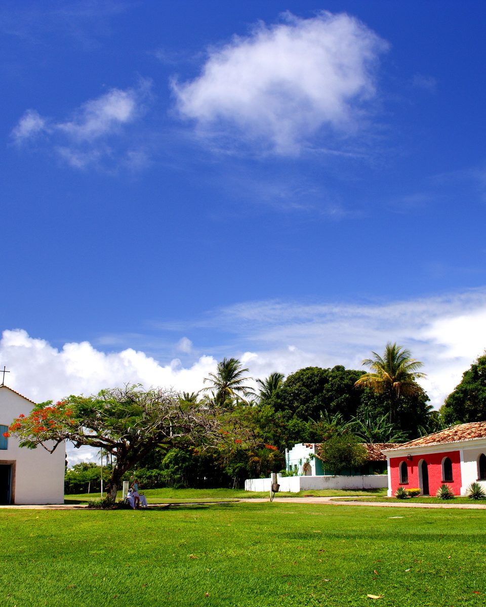 Quadrado de Trancoso num dia de céu azul, com a igreja branca de São João Batista, o gramado verde e as casinhas coloridas de telhado de barro sob as árvores.