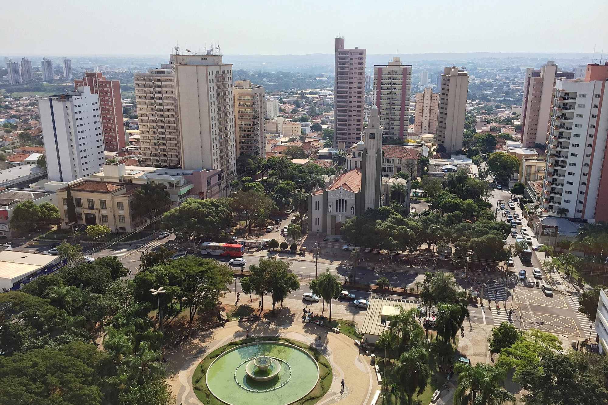 Vista do alto do Centro de Presidente Prudente, com o chafariz da Praça 9 de Julho em primeiro plano, a Catedral de São Sebastião no meio das árvores e prédios residenciais ao fundo, num dia de sol.