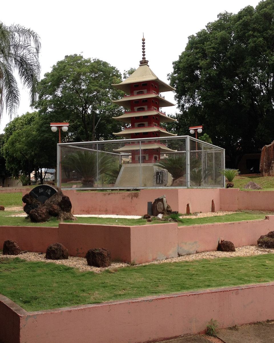 Pagode vermelho de estilo japonês numa praça ajardinada de Presidente Prudente, com palmeiras, pedras e árvores em volta.