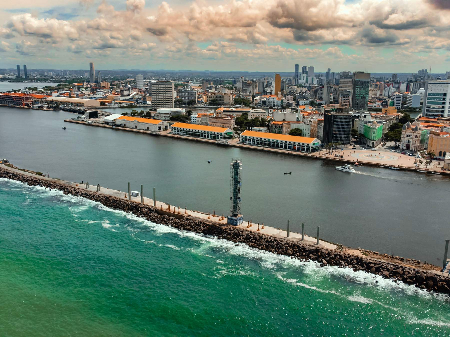 Molhe dos arrecifes em frente ao Recife visto do alto, com a Torre de Cristal do Parque das Esculturas Francisco Brennand, o mar verde de um lado, as águas do porto do outro e o Bairro do Recife com prédios ao fundo, sob nuvens de fim de tarde.