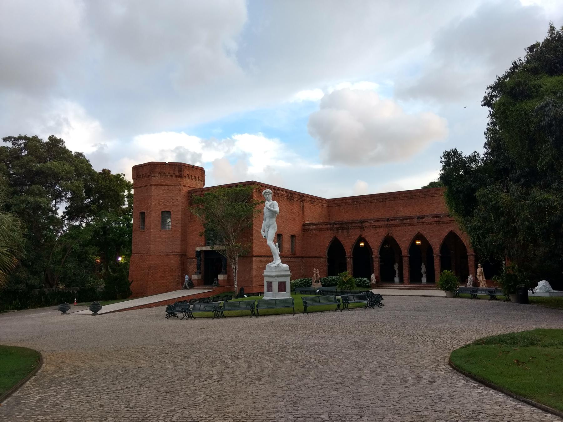 Castelo de tijolos do Instituto Ricardo Brennand, na Várzea, com torre, arcadas, uma estátua branca no pátio circular, gramado e mata ao redor sob céu com nuvens.