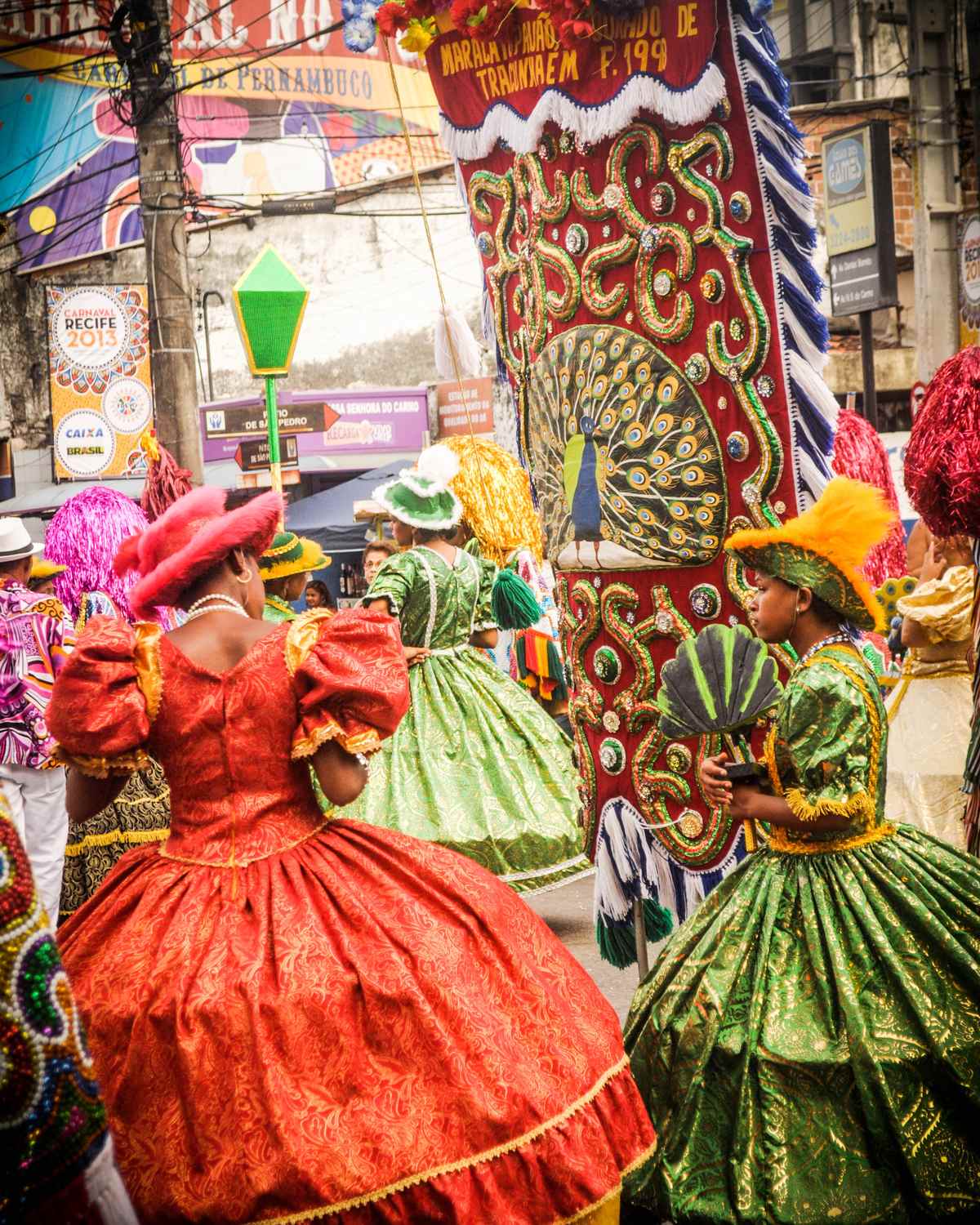 Damas de maracatu com vestidos rodados vermelho e verde, chapéus e adereços coloridos, desfilando entre estandartes bordados numa rua do centro do Recife durante o Carnaval.