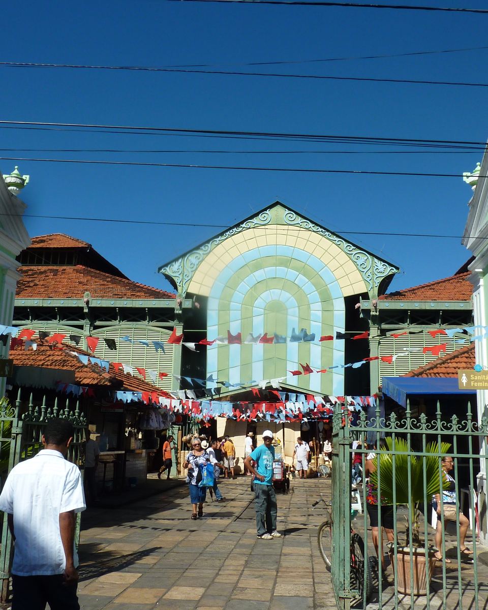 Entrada do Mercado de São José, no Recife, com a fachada de ferro em arco e bandeirinhas coloridas, pessoas passando pela calçada e casarões verde e branco dos dois lados, sob céu azul.
