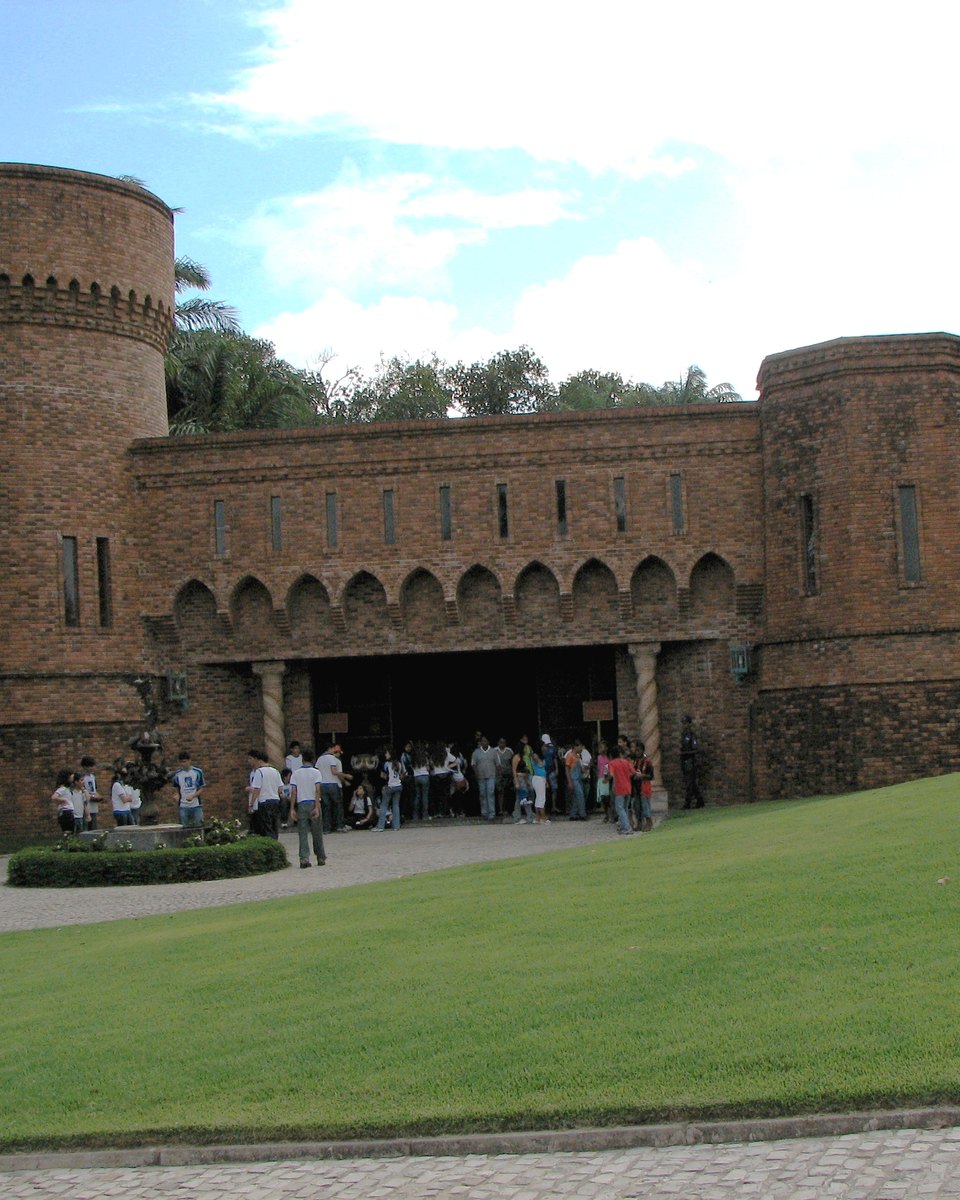 Castelo São João, de tijolos e torres redondas, no Instituto Ricardo Brennand, na Várzea, com um grupo de visitantes na entrada em arco, gramado verde em primeiro plano e árvores ao redor.