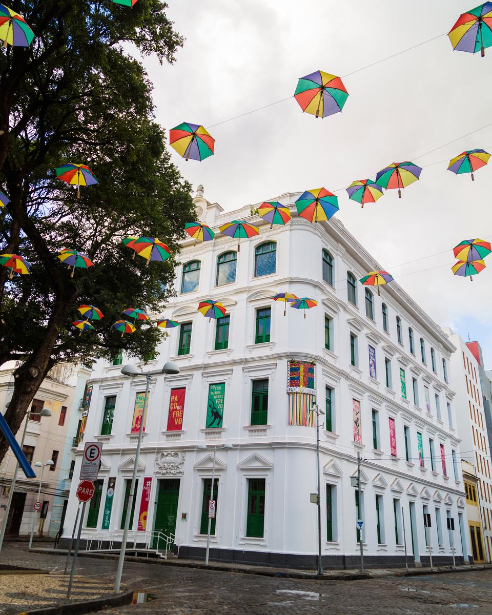 Fachada branca do Paço do Frevo, no Bairro do Recife, com janelas verdes e vermelhas, sombrinhas de frevo coloridas penduradas em fios sobre a rua e uma árvore à esquerda, sob céu claro.