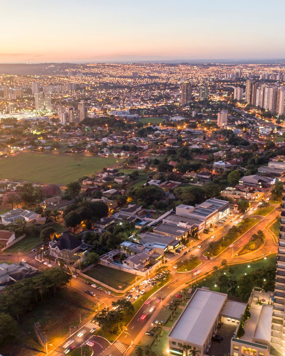Vista aérea da Avenida Professor João Fiúza, em Ribeirão Preto, ao entardecer, com prédios altos, casas e as luzes dos carros na avenida.