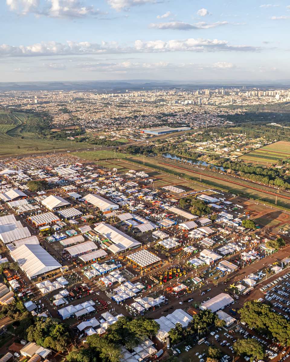 Vista aérea da Agrishow ao entardecer, com pavilhões brancos, estacionamento lotado e plantações em volta, e os prédios de Ribeirão Preto ao fundo.