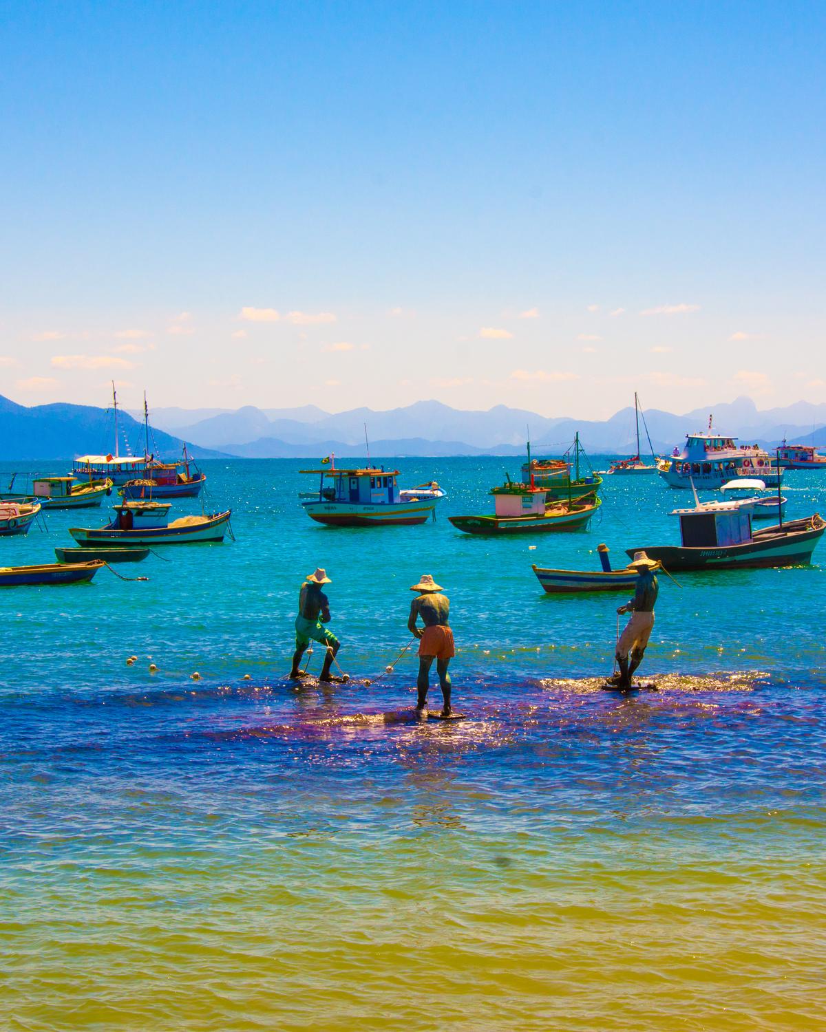 Estátuas de três pescadores na água rasa e barcos coloridos ancorados numa praia de Búzios, com montanhas ao fundo.