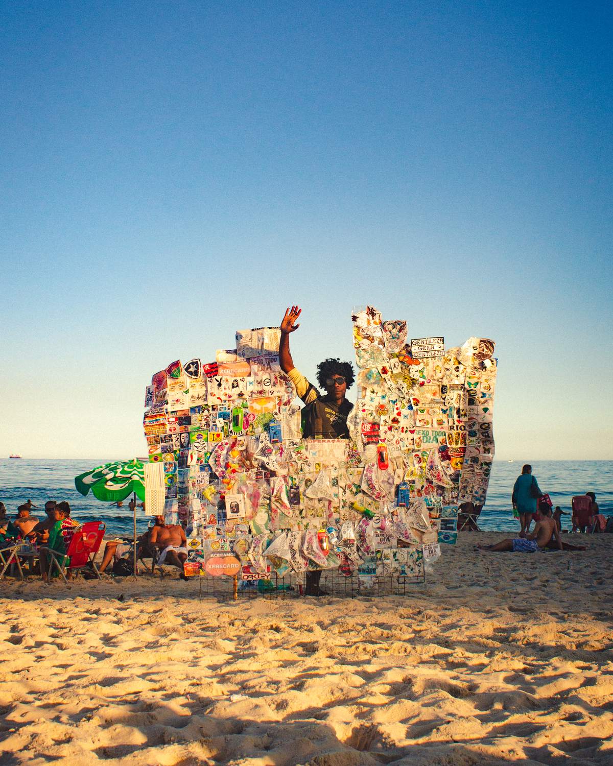 Vendedor acenando atrás de um painel coberto de adesivos com gírias e memes cariocas, na areia da praia de Copacabana, com banhistas ao fundo.