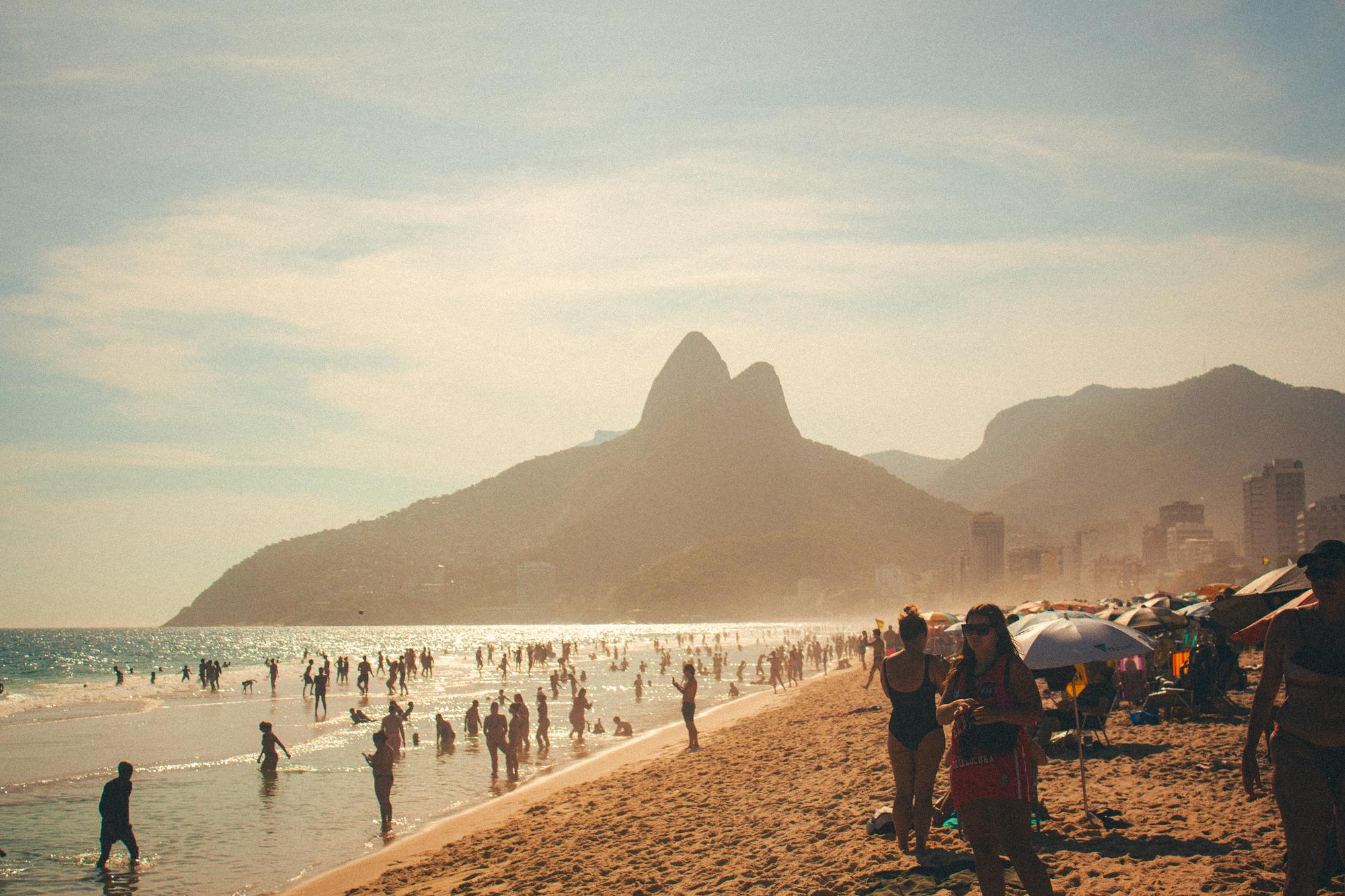 Banhistas na areia e na água da praia de Ipanema numa tarde de sol, com o Morro Dois Irmãos e os prédios do Leblon ao fundo.