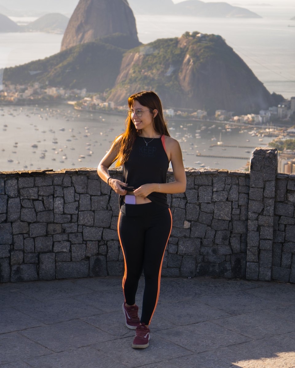 Mulher de roupa de treino sorrindo no Mirante Dona Marta, com o Pão de Açúcar, a Baía de Guanabara e barcos ao fundo, numa manhã de sol.