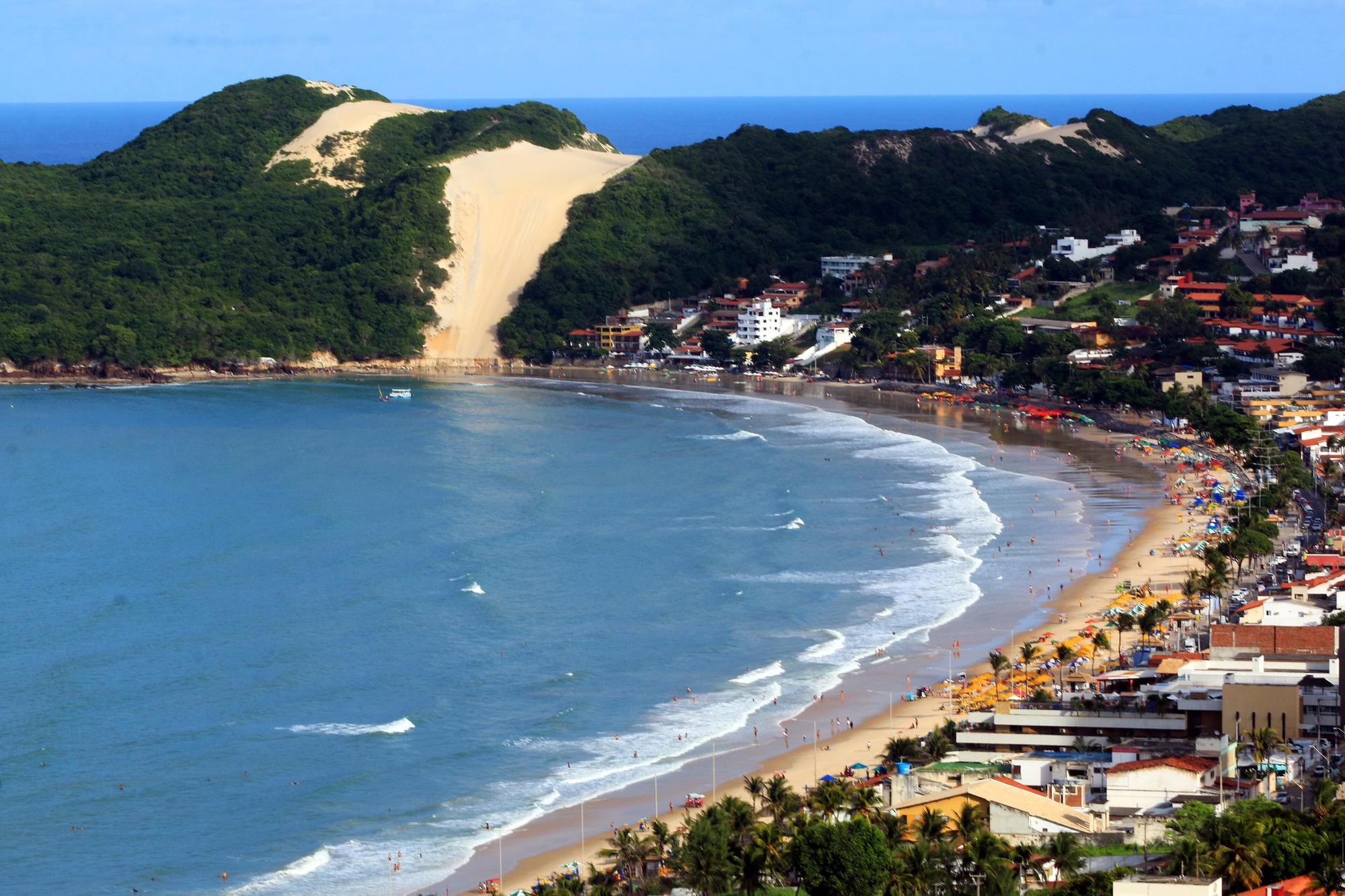 Praia de Ponta Negra, em Natal, com a faixa de areia, o mar esverdeado e o Morro do Careca, a duna de areia clara cercada de vegetação, no fim da praia