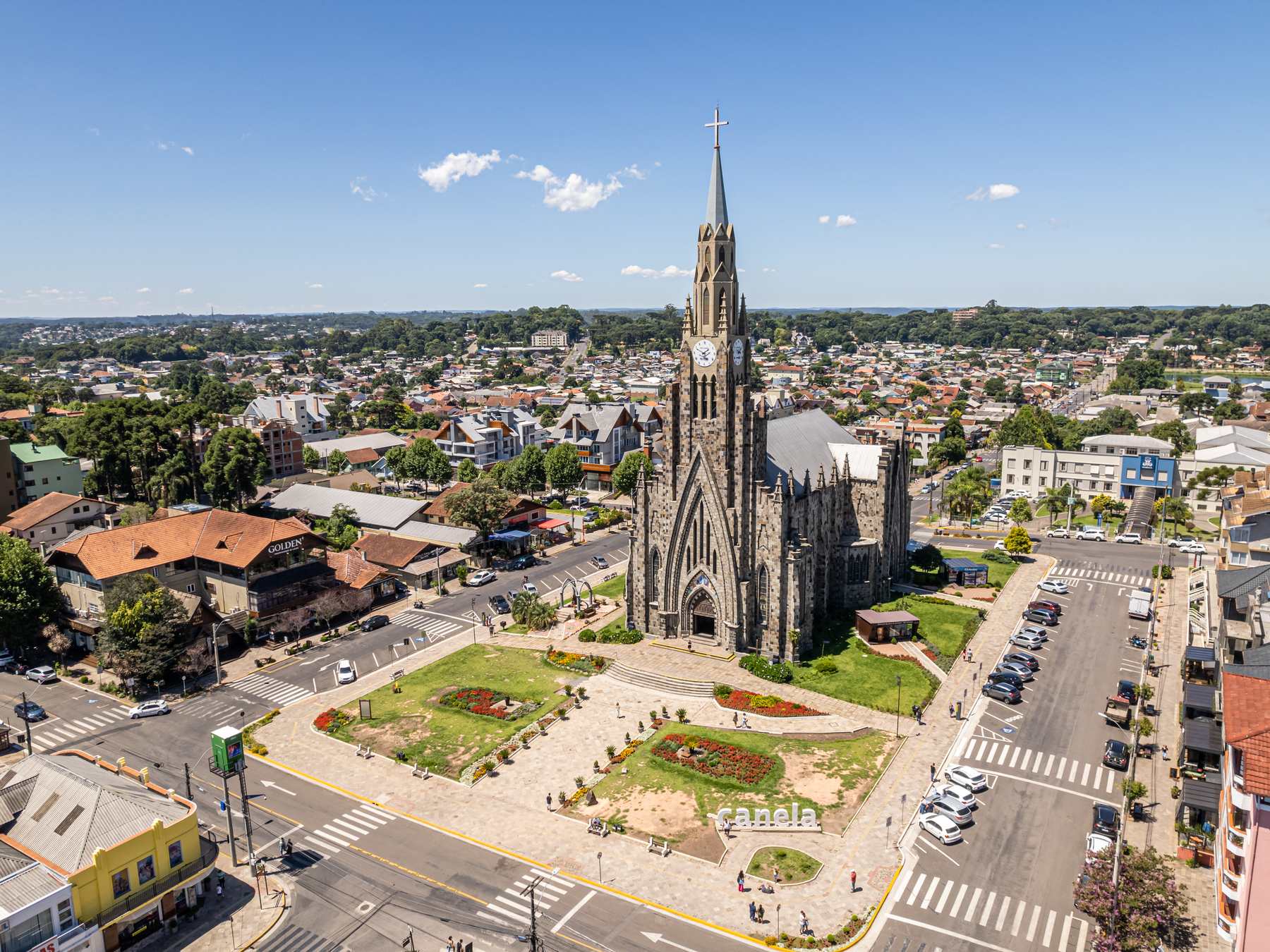 Vista do alto do centro de Canela, com a Catedral de Pedra de torre pontiaguda, a praça ajardinada à frente e a cidade se estendendo até o horizonte.