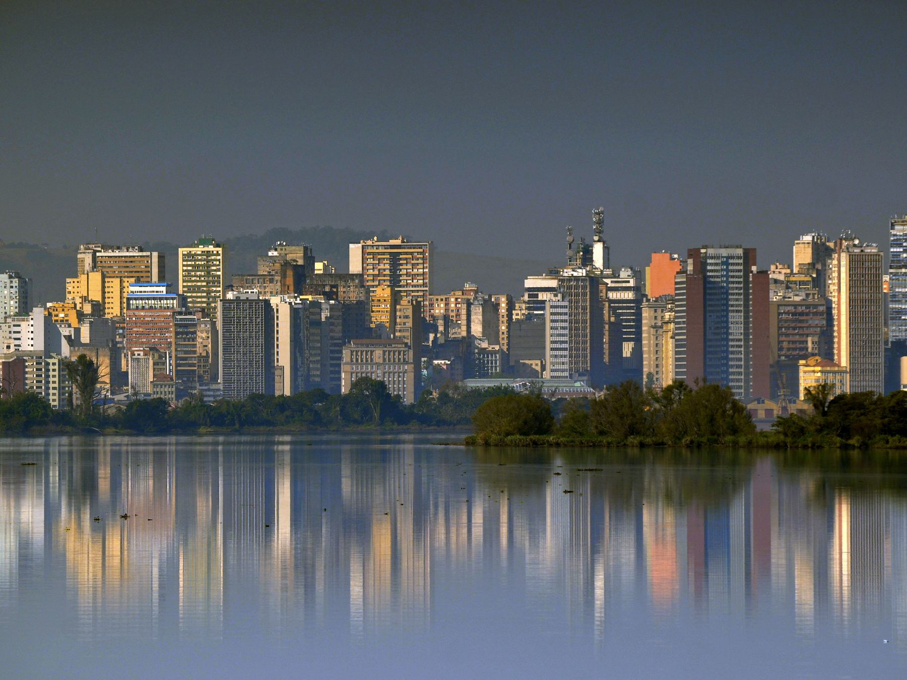 Prédios de Porto Alegre refletidos na água parada do Guaíba no fim da tarde, com a vegetação da margem na linha d'água.