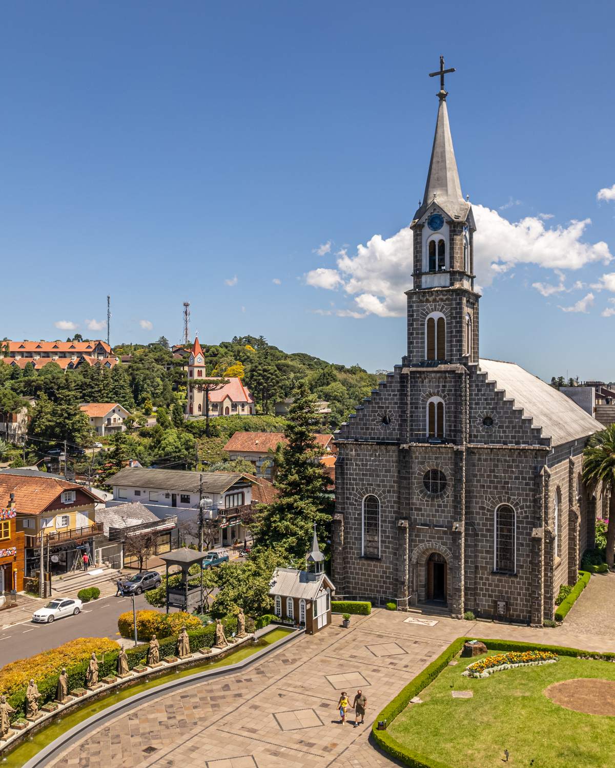 Igreja Matriz São Pedro, de pedra, com a praça ajardinada em frente e o centro de Gramado em volta, vistos do alto num dia de céu azul.
