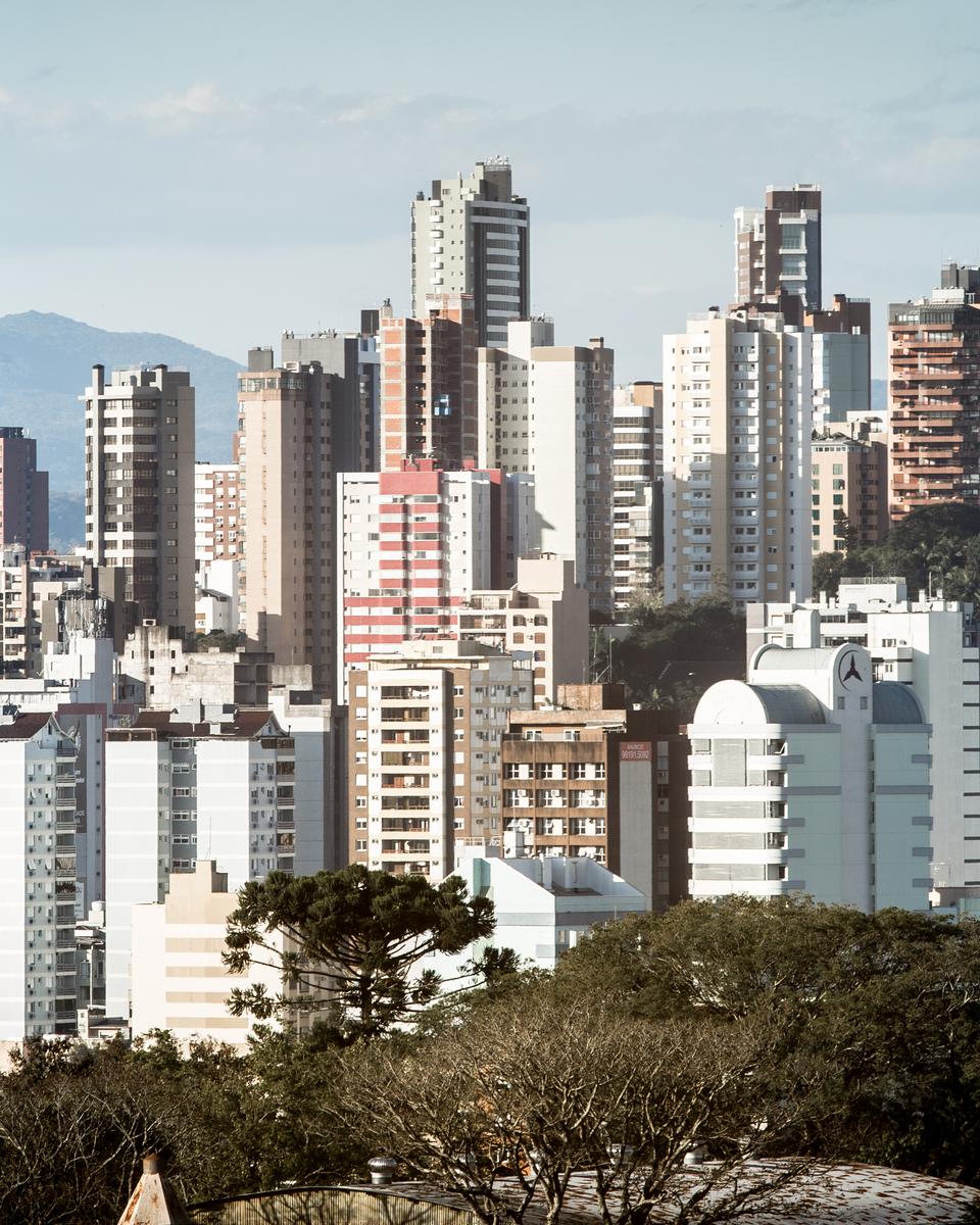 Prédios do centro de Novo Hamburgo com a serra ao fundo e a copa das árvores em primeiro plano.