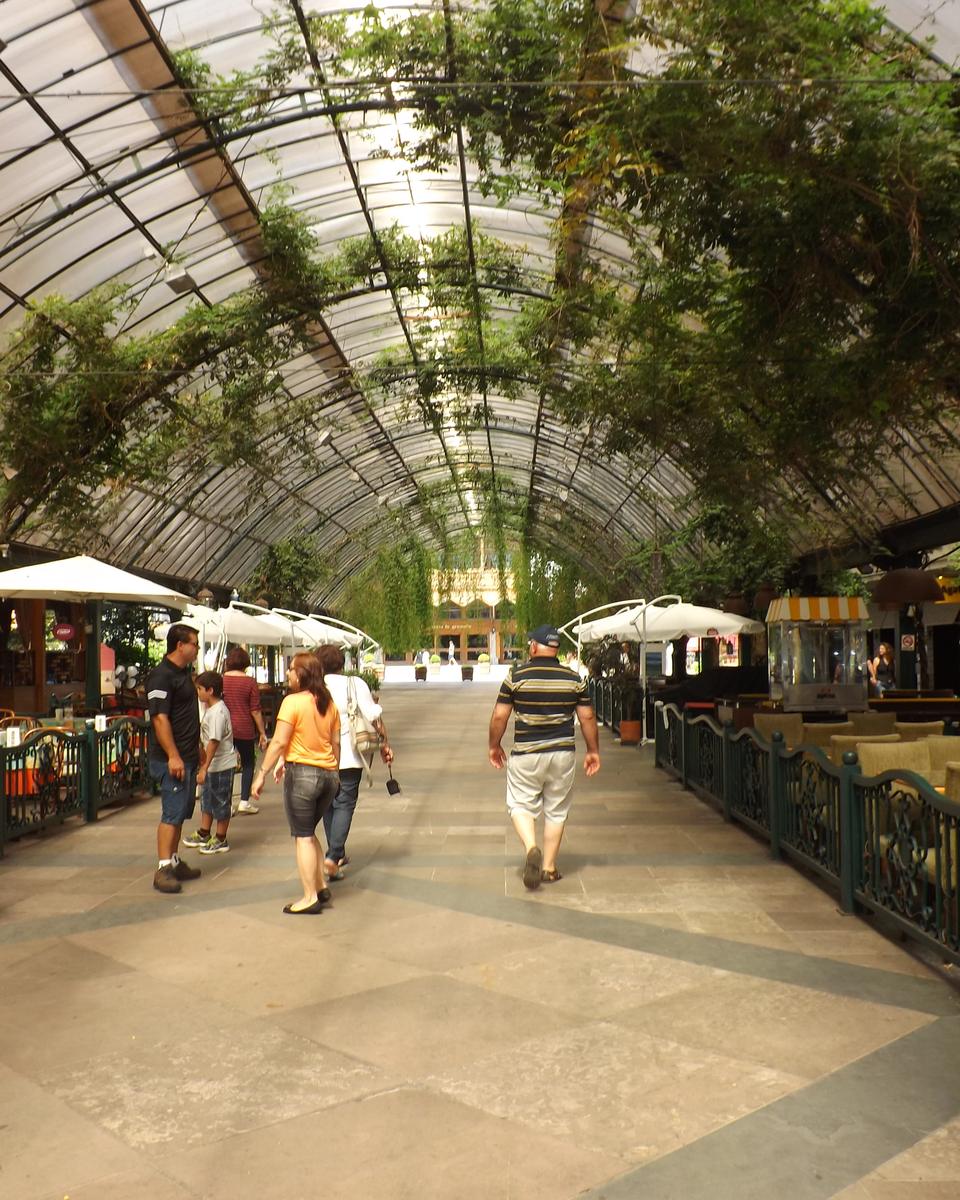 Rua Coberta de Gramado, com teto de vidro, plantas penduradas, mesas de restaurante dos dois lados e gente passeando pelo calçamento.