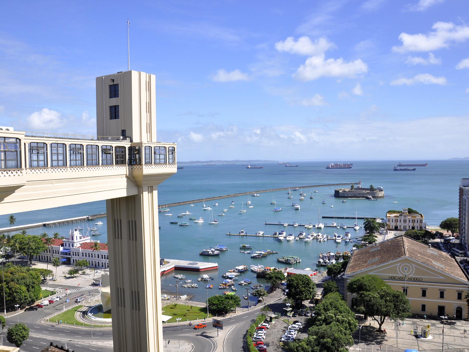 Elevador Lacerda visto do alto da Cidade Alta, em Salvador, com a passarela em primeiro plano, o Mercado Modelo, a marina cheia de barcos e a Baía de Todos-os-Santos azul ao fundo.