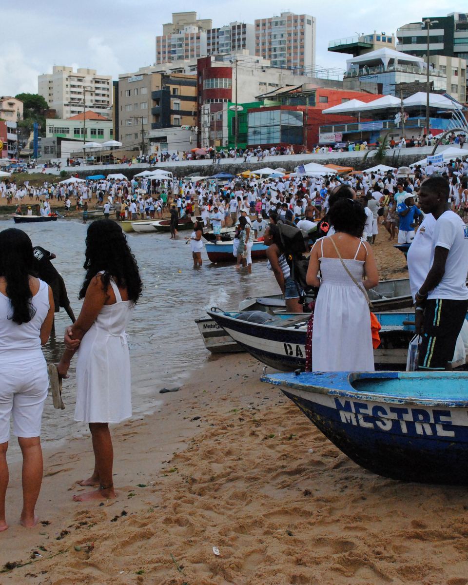 Pessoas vestidas de branco caminhando na areia do Rio Vermelho, em Salvador, entre barcos de pesca coloridos encalhados na praia, durante a Festa de Iemanjá, com os prédios e casas do bairro ao fundo.