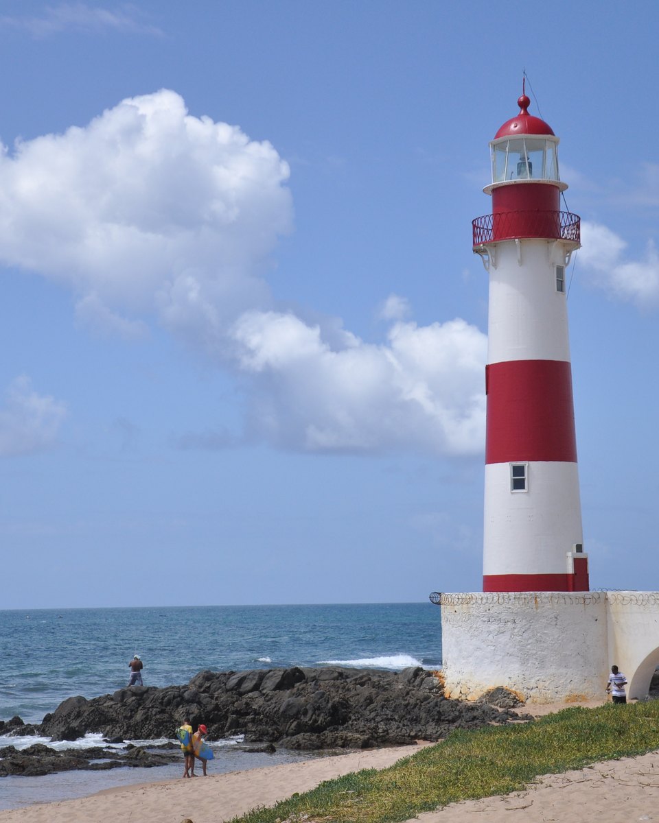 Farol de Itapuã, listrado de vermelho e branco, sobre a mureta branca à beira das pedras da praia, em Salvador, com pessoas na areia, o mar azul e nuvens brancas no céu.