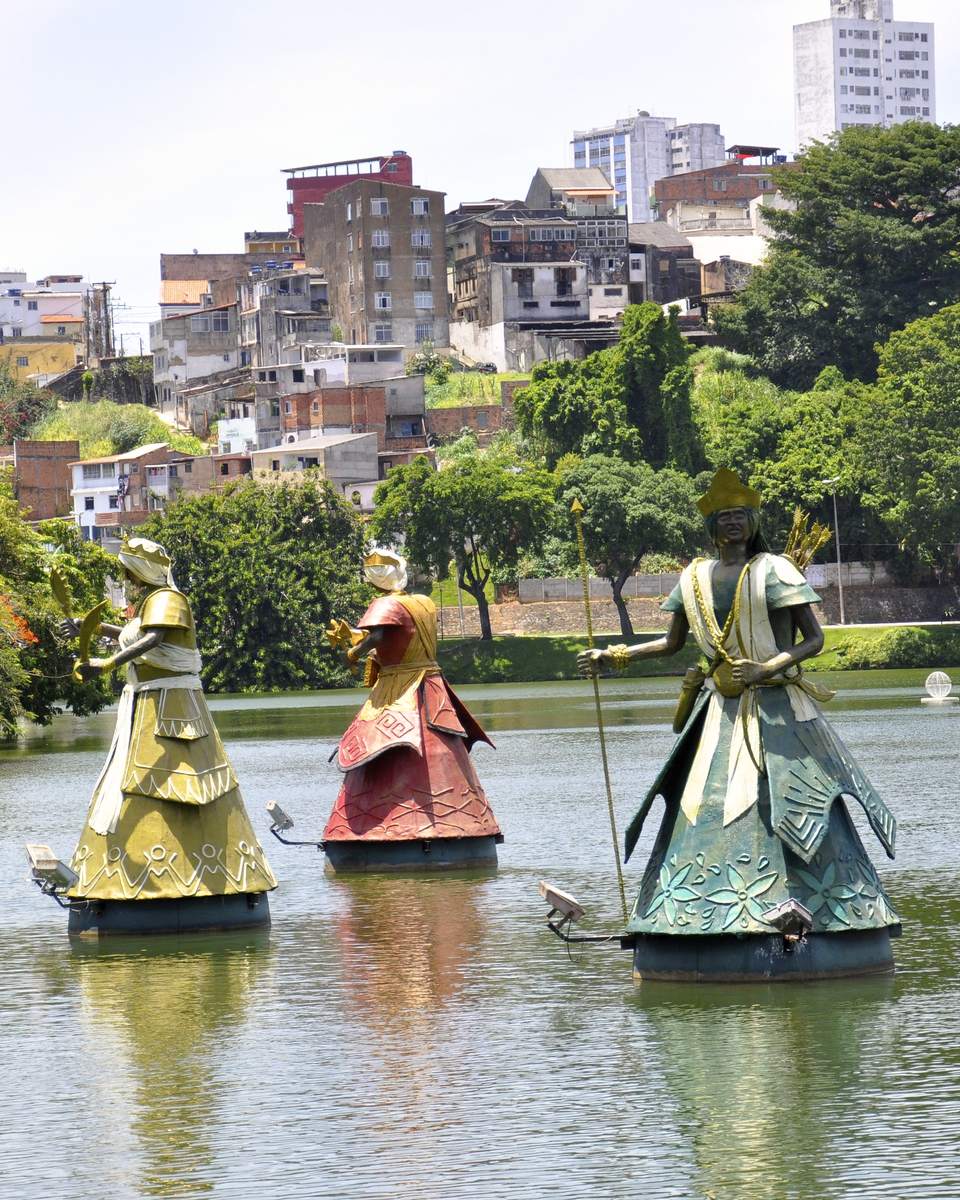 Esculturas gigantes dos orixás, com saias coloridas e metálicas, sobre a água do Dique do Tororó, em Salvador, com árvores na margem e os prédios e casas do bairro ao fundo, num dia claro.