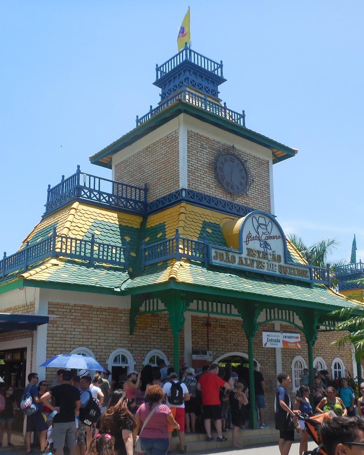 Visitantes em fila na entrada do Beto Carrero World, em Penha, diante da estação de torre com relógio, telhado verde e amarelo e grades azuis, num dia de sol.