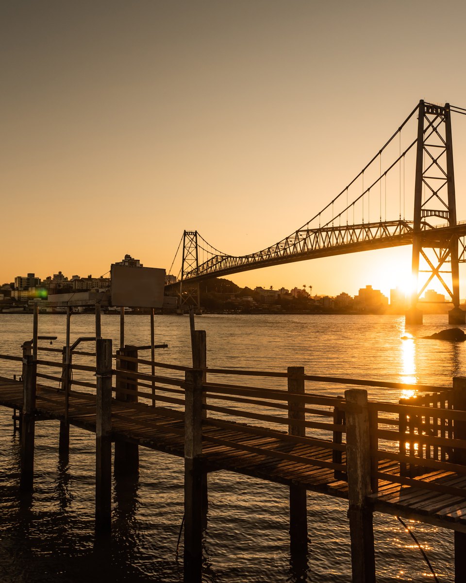 Pôr do sol atrás da Ponte Hercílio Luz, em Florianópolis, com um trapiche de madeira em primeiro plano e o reflexo do sol na água da baía.