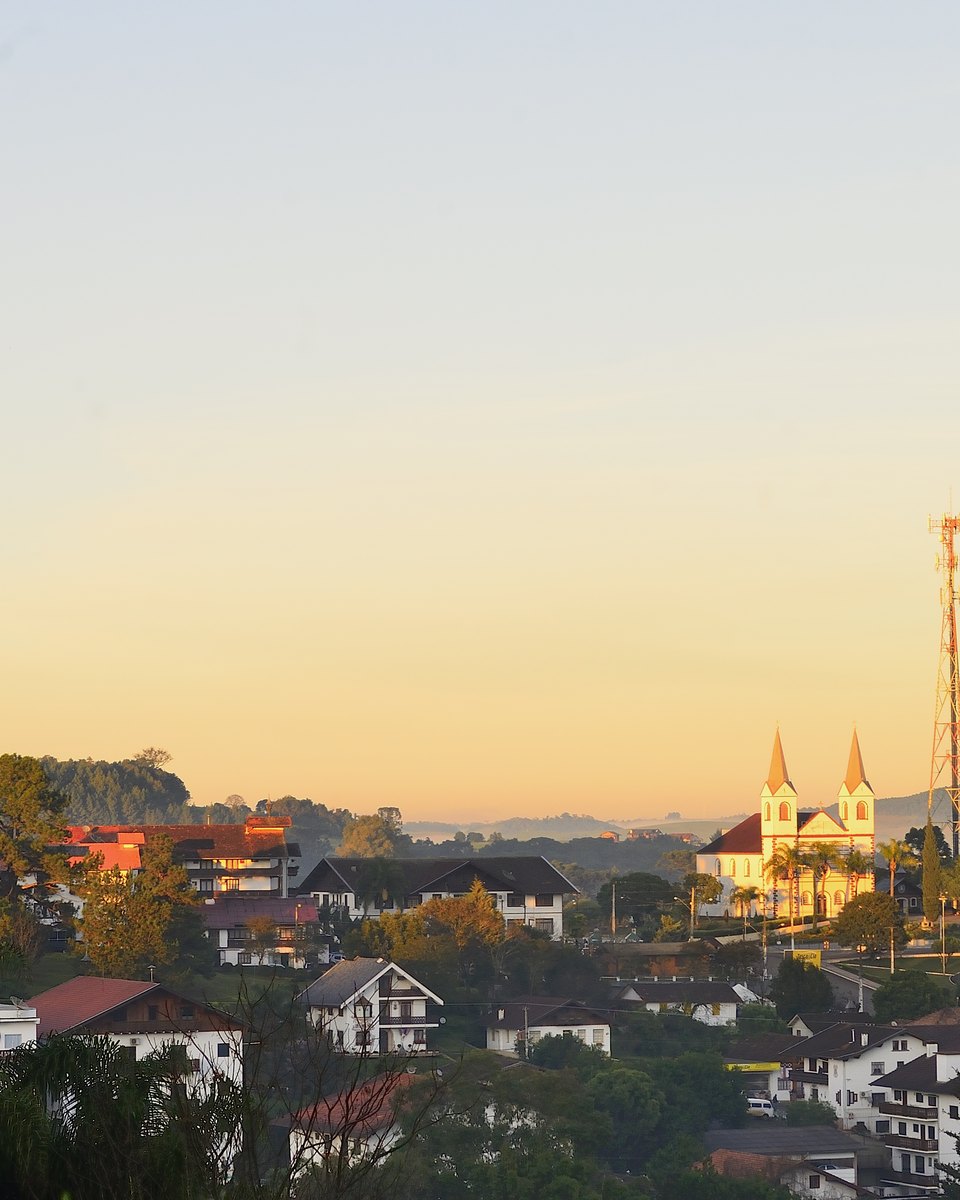 Treze Tílias vista do alto ao amanhecer, com casas de estilo tirolês entre as árvores, a igreja de duas torres ao fundo e o céu dourado.