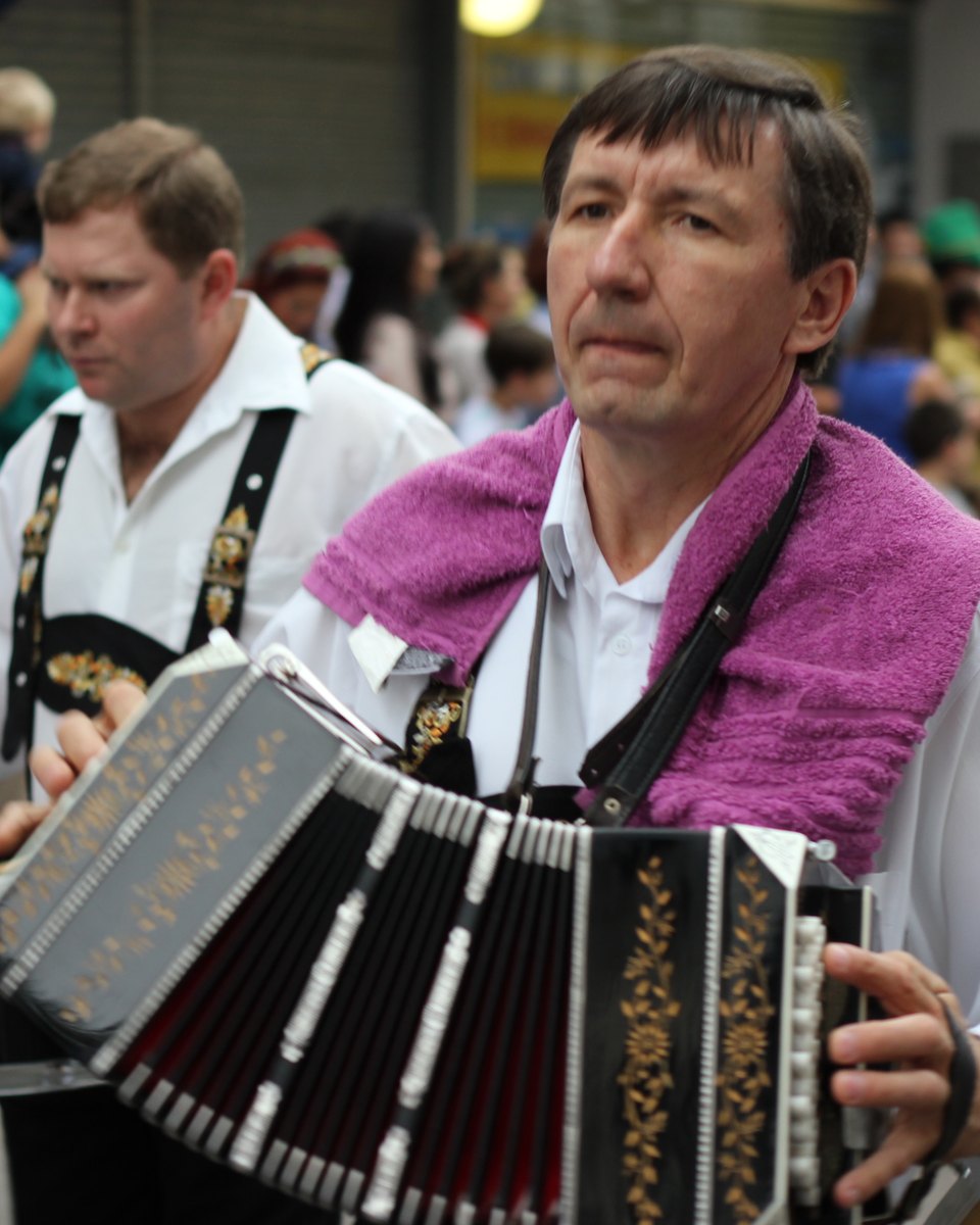 Músico de camisa branca, suspensórios e toalha lilás no pescoço tocando bandoneon no desfile da Oktoberfest de Blumenau, ao lado de outros músicos da banda e do público.
