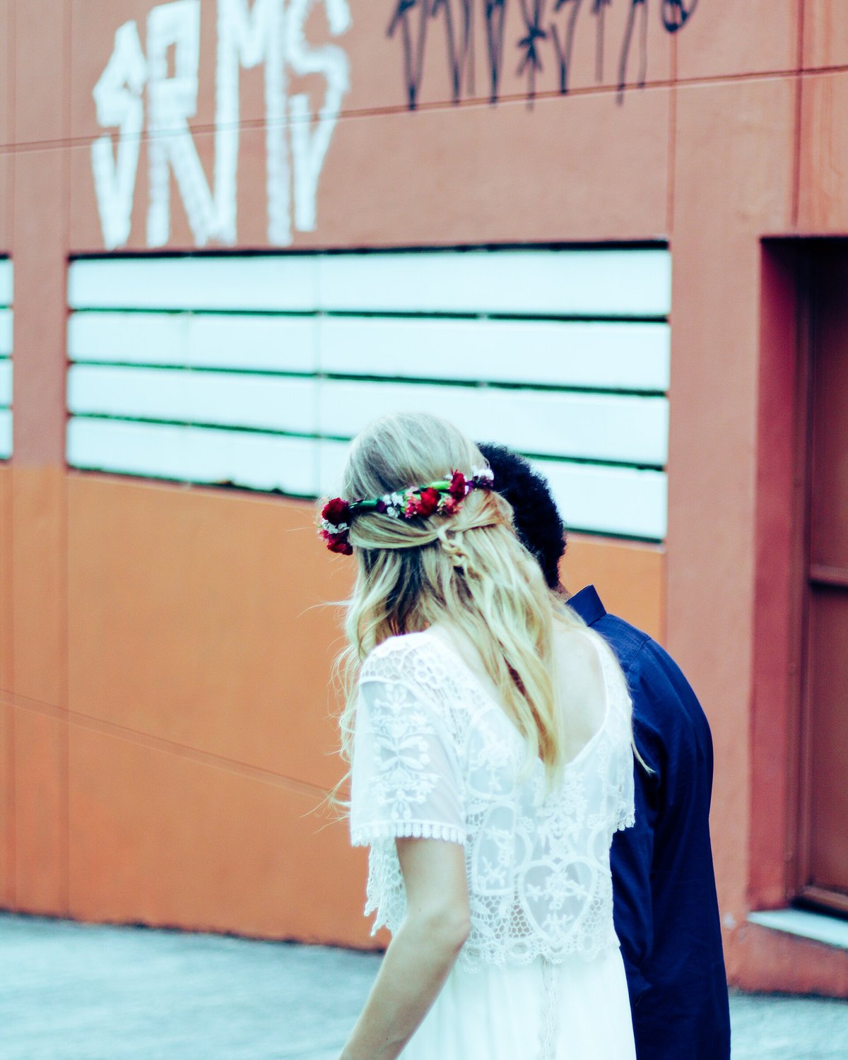 Casal de noivos caminhando por uma rua de Santo André, ela de vestido branco rendado e coroa de flores, ele de terno azul, diante de um muro laranja.