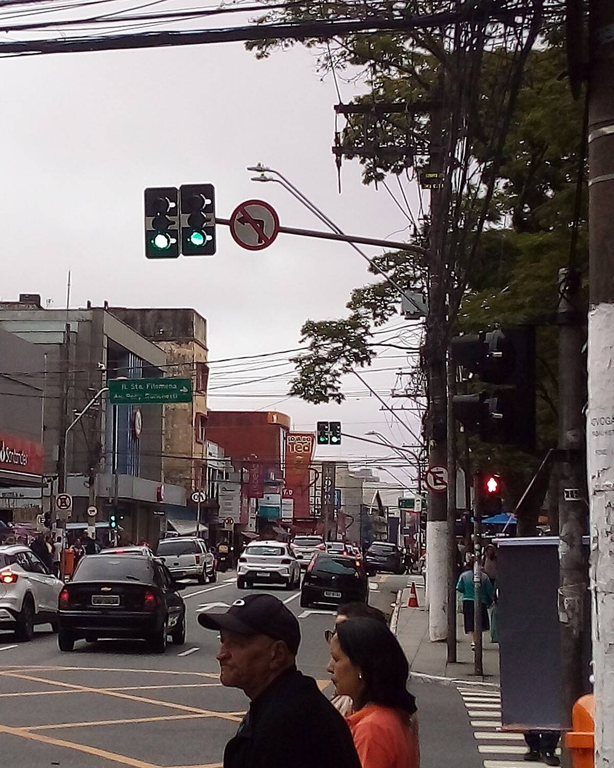 Homem de boné e mulher de blusa laranja esperando para atravessar a Rua Marechal Deodoro, no Centro de São Bernardo do Campo, com lojas, semáforos e carros ao fundo.