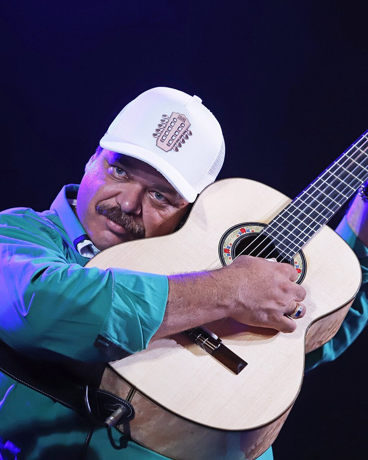 Violeiro de boné branco e camisa verde abraçado à viola caipira, sorrindo no palco do Festival Nacional da Moda de Viola, em São José do Rio Preto.