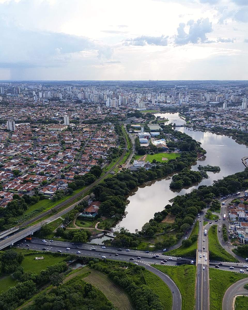 Vista aérea de São José do Rio Preto com bairros de casas, o trevo de uma rodovia, a Represa Municipal serpenteando entre áreas verdes e os prédios do centro no horizonte.