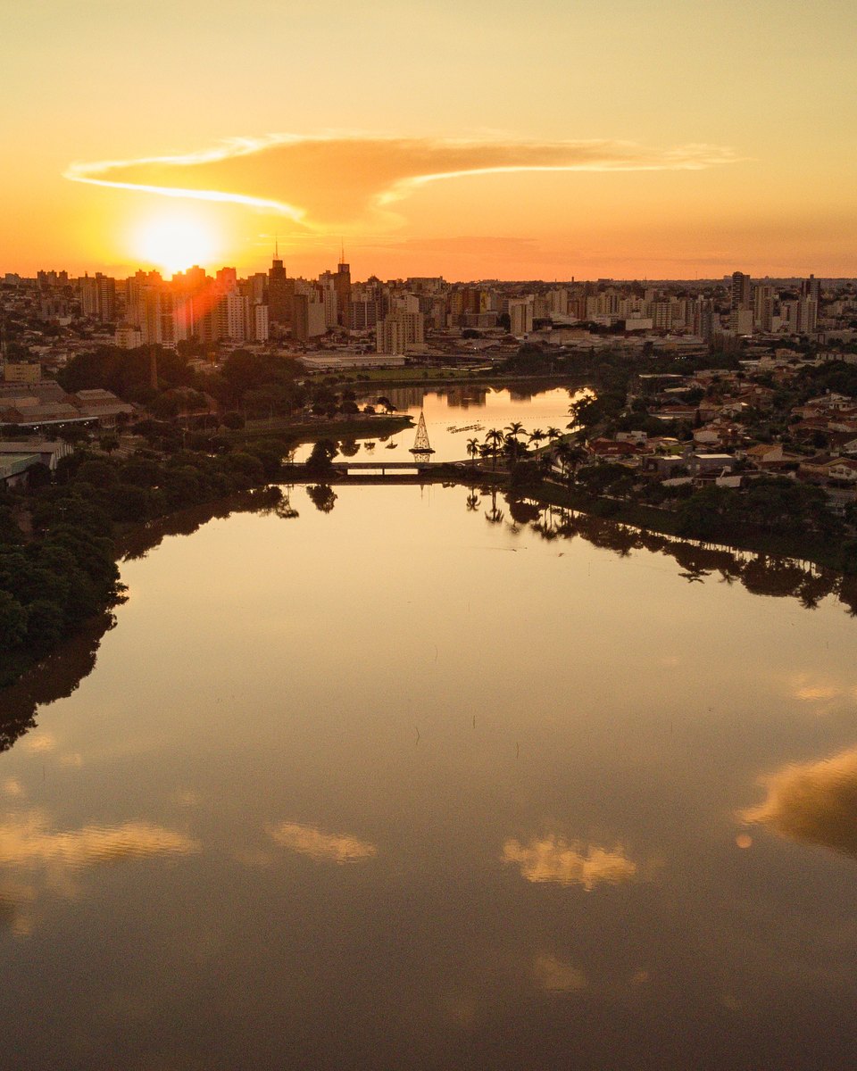 Represa Municipal de São José do Rio Preto vista de drone ao pôr do sol, com o céu laranja refletido na água, margens arborizadas e os prédios da cidade no horizonte.
