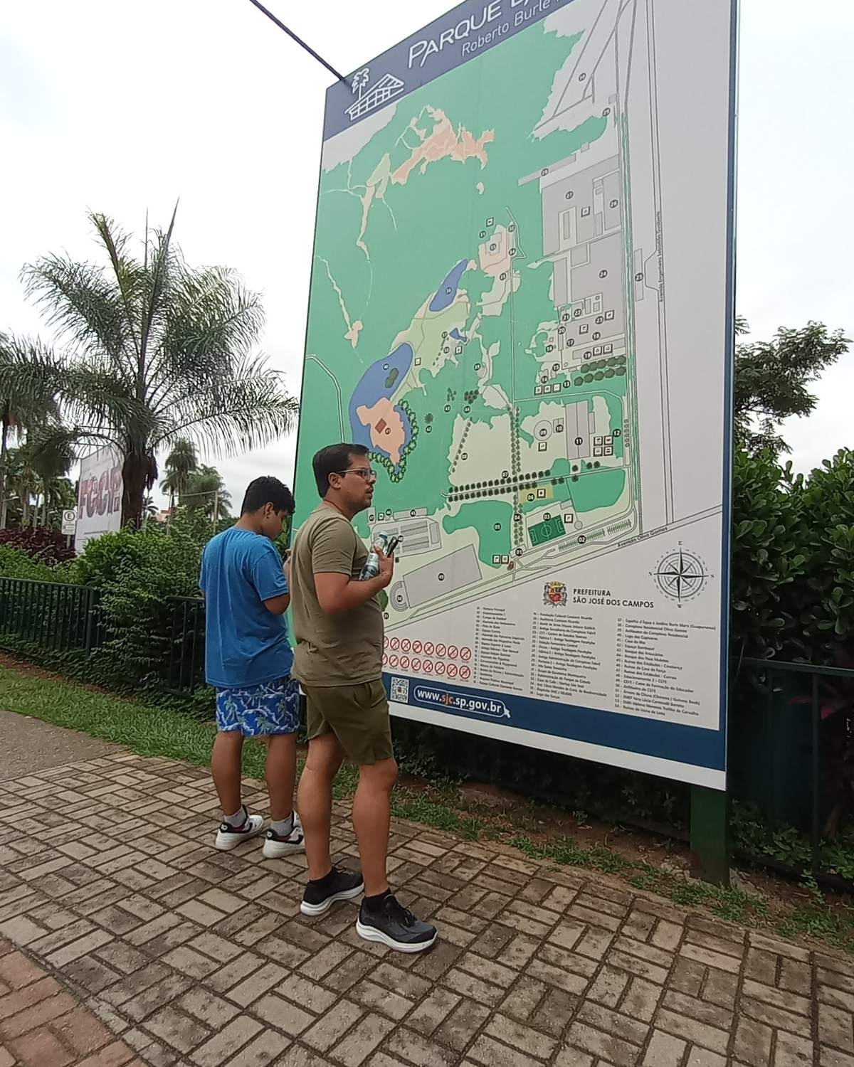 Dois rapazes consultando o mapa na entrada do Parque da Cidade Roberto Burle Marx, em São José dos Campos, com palmeiras ao fundo.