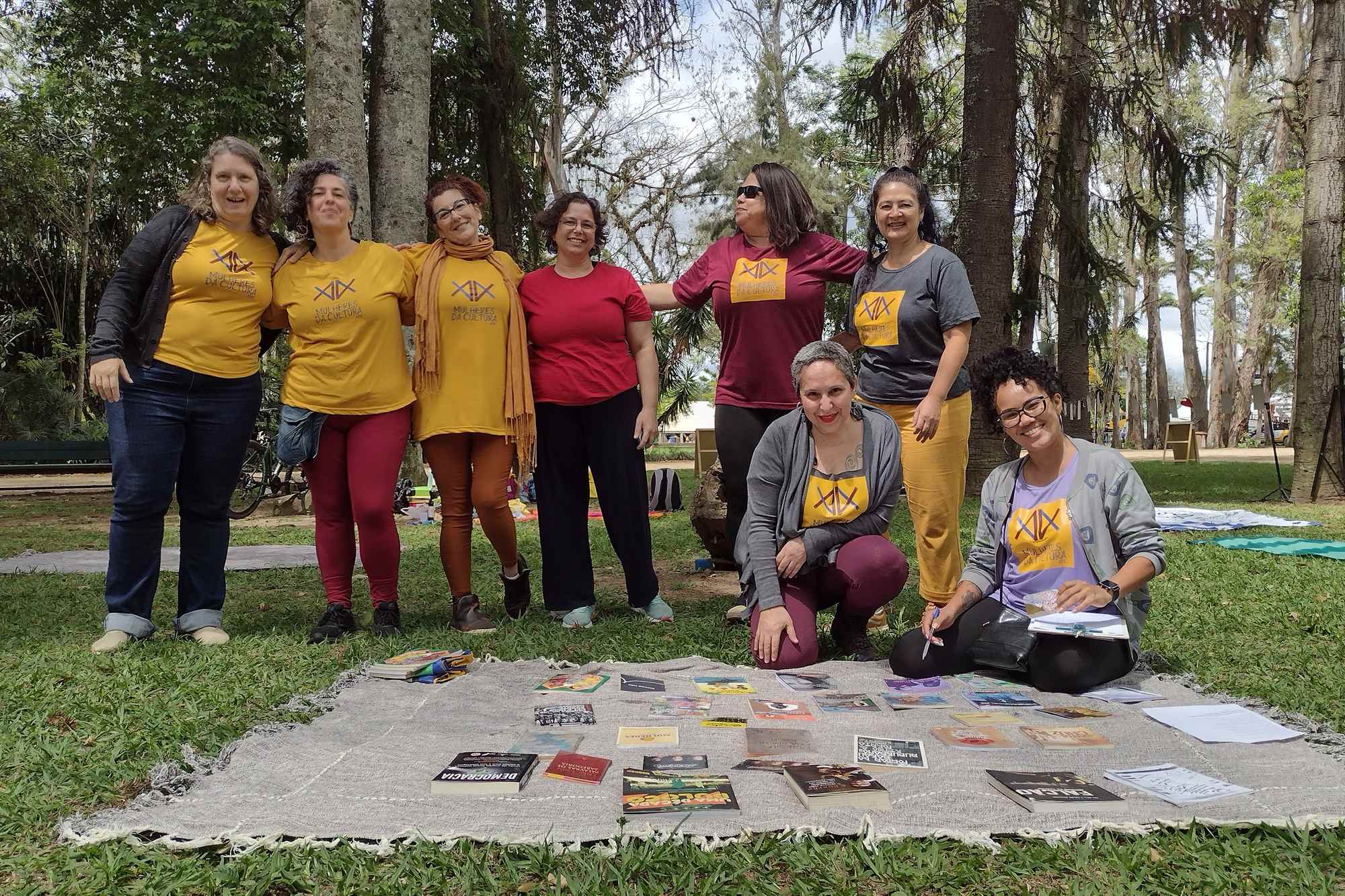 Grupo de mulheres de camiseta amarela e vermelha sorrindo sob as árvores do Parque da Cidade, em São José dos Campos, com livros espalhados numa manta sobre a grama.