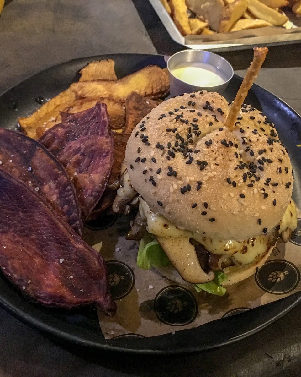 Hambúrguer com gergelim preto e batata-doce roxa frita num prato escuro, na mesa de um bar de São José dos Campos.