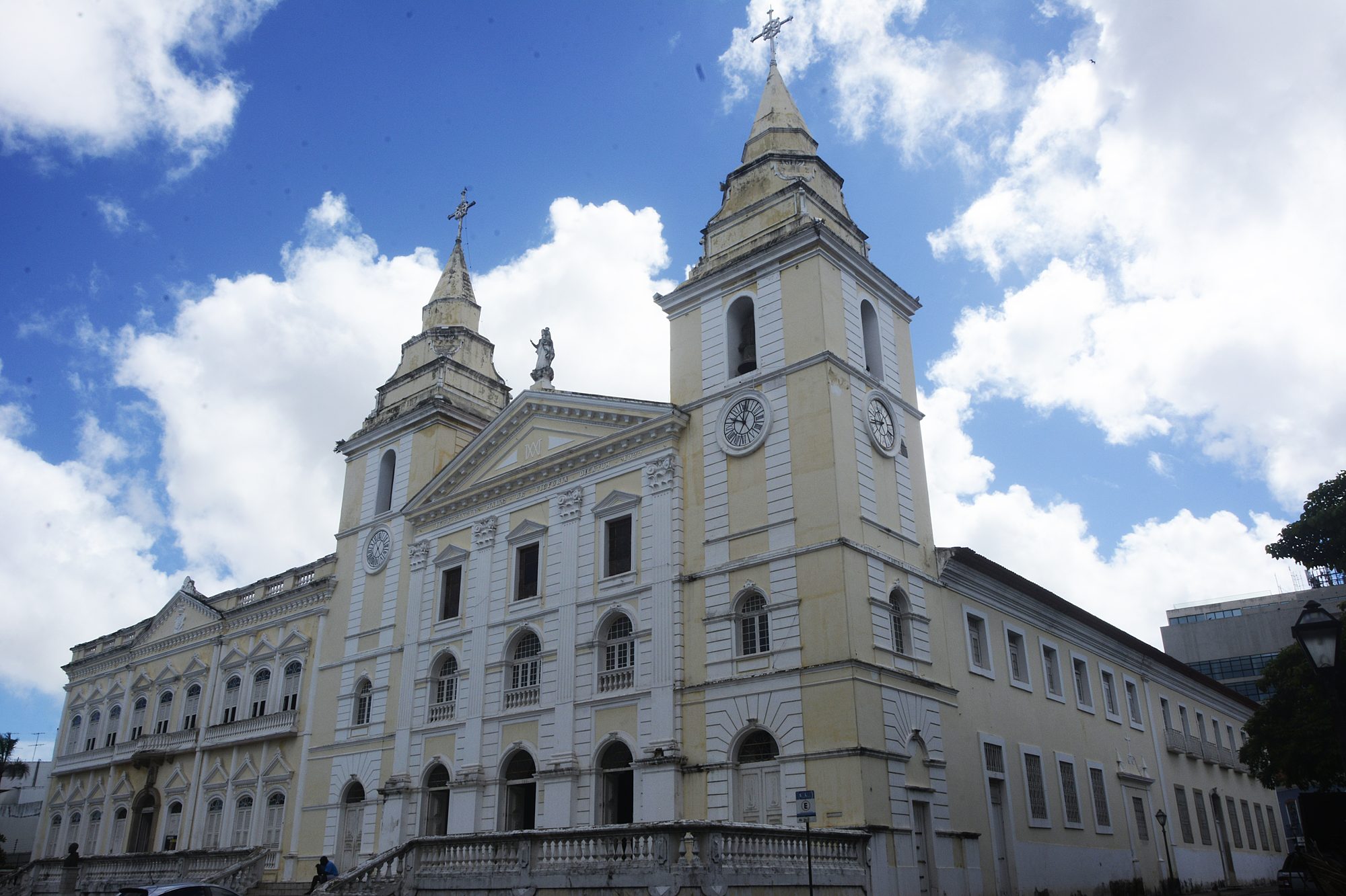 Fachada da Catedral da Sé de São Luís vista de baixo, com as duas torres, o relógio e o frontão recortados no céu azul com nuvens
