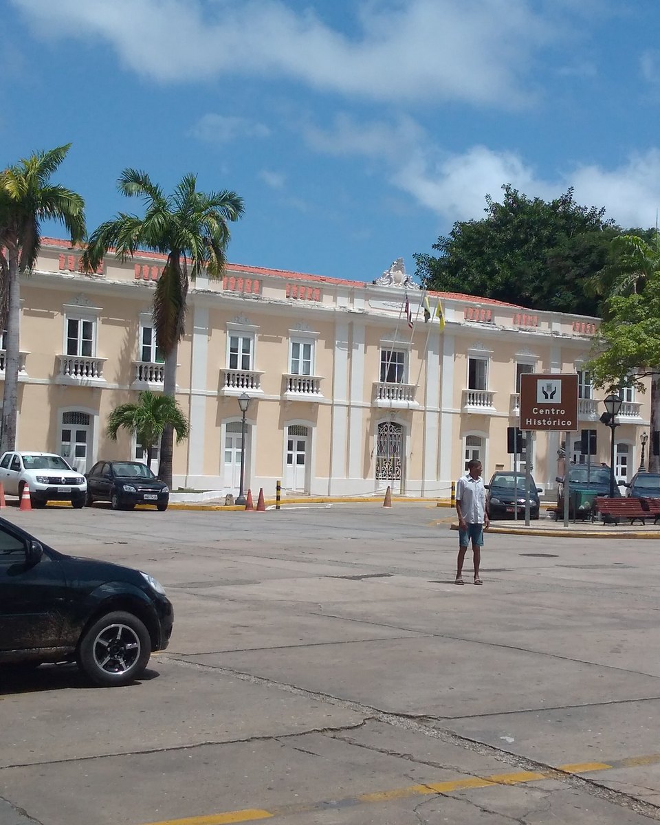 Fachada bege do Palácio La Ravardière com palmeiras na frente e pessoas atravessando a praça em um dia de sol