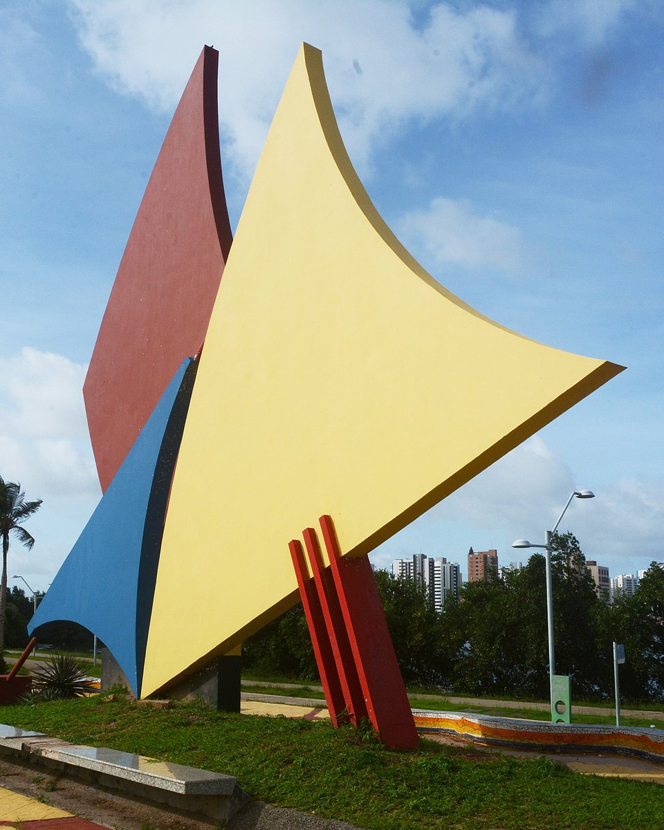 Escultura colorida em forma de vela no parque da Lagoa da Jansen, vista do calçadão, com a lagoa e os prédios da cidade ao fundo