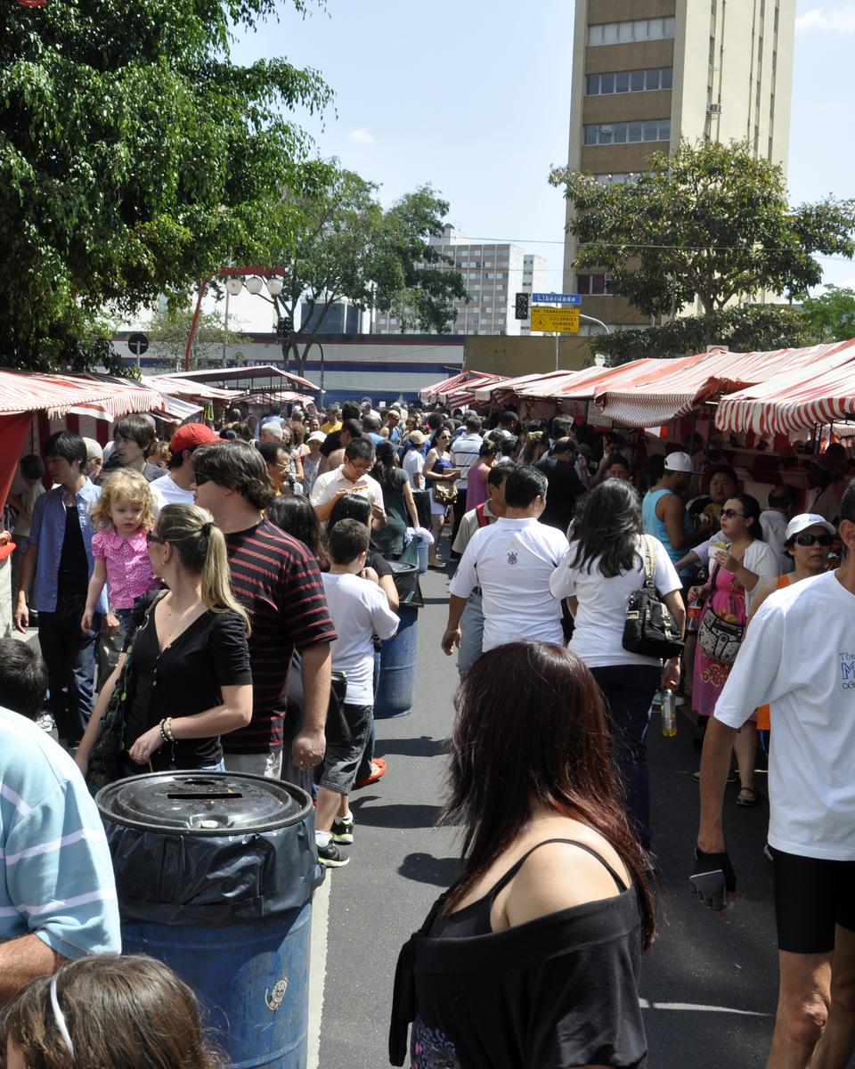 Pessoas circulando entre barracas listradas de vermelho e branco na feira de fim de semana da Liberdade.