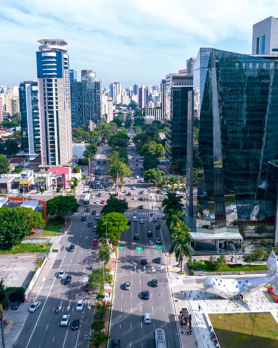 Vista do alto da Avenida Brigadeiro Faria Lima, no Itaim Bibi, com prédios comerciais de vidro, canteiro arborizado e carros nas pistas, num dia de sol em São Paulo.