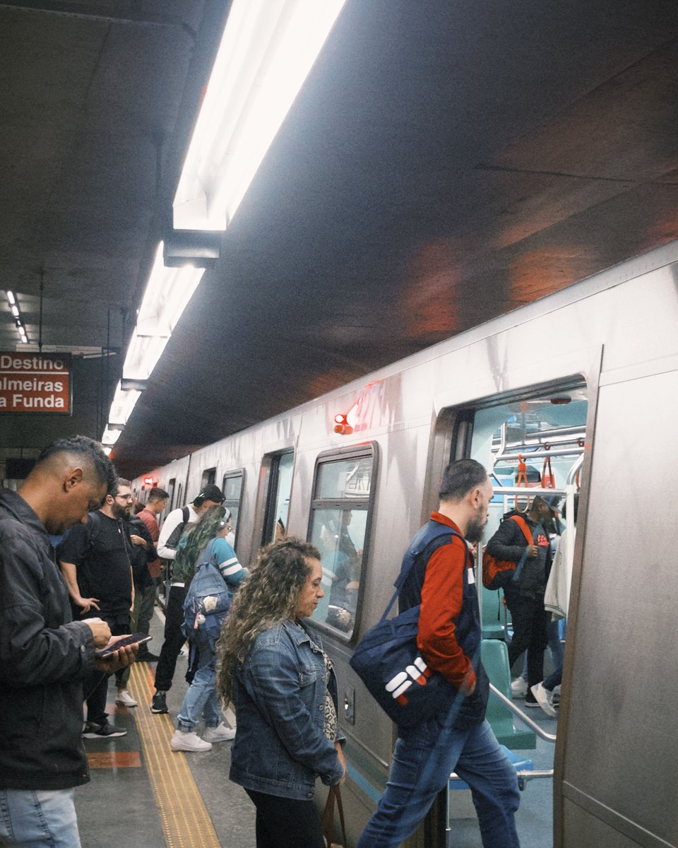 Passageiros embarcando num trem do Metrô de São Paulo na plataforma de uma estação da Linha 3-Vermelha, com um homem olhando o celular e a placa de destino Palmeiras-Barra Funda ao fundo.