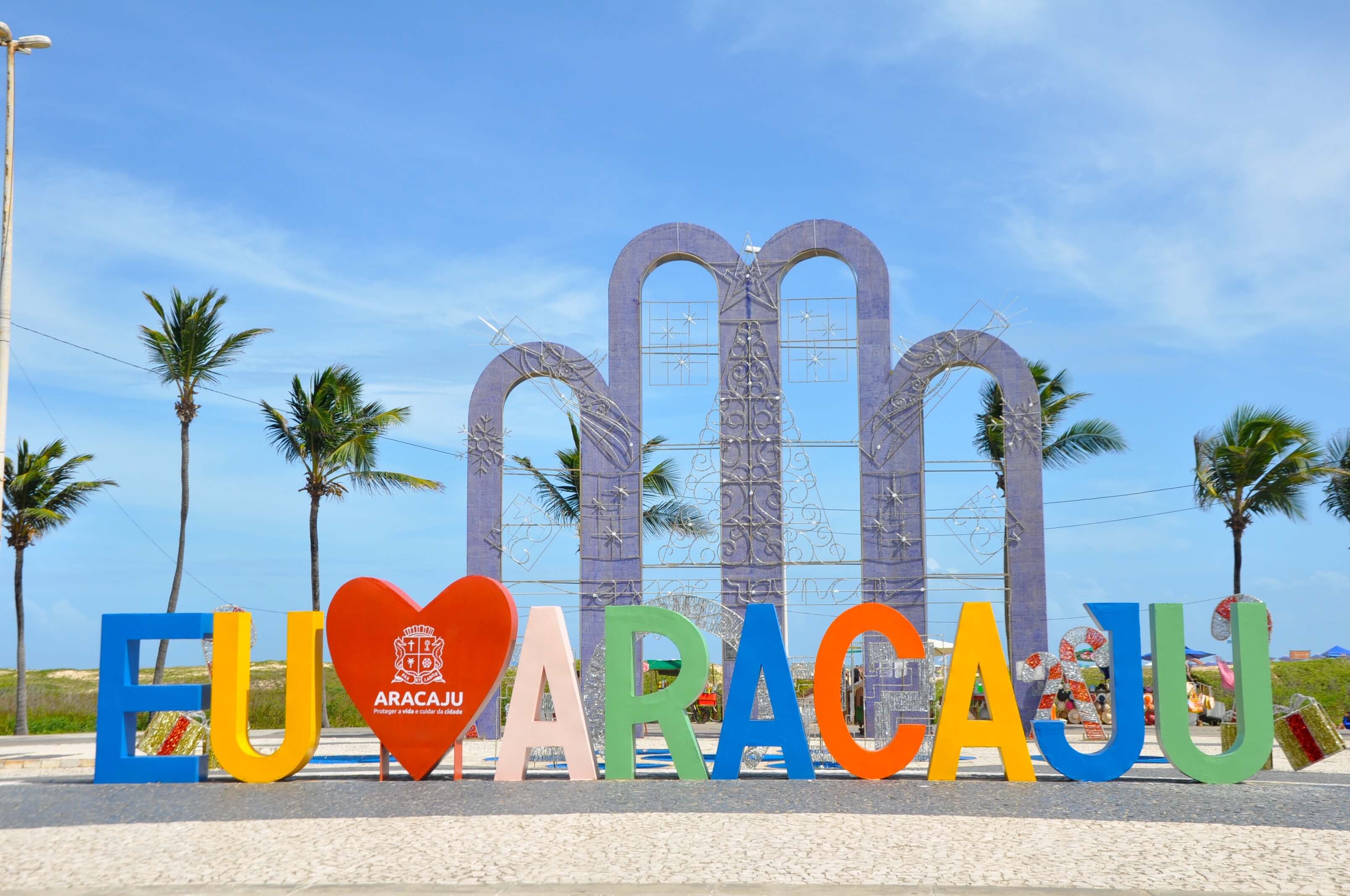 Letreiro colorido "Eu Amo Aracaju" com coqueiros ao fundo, na orla da capital sergipana