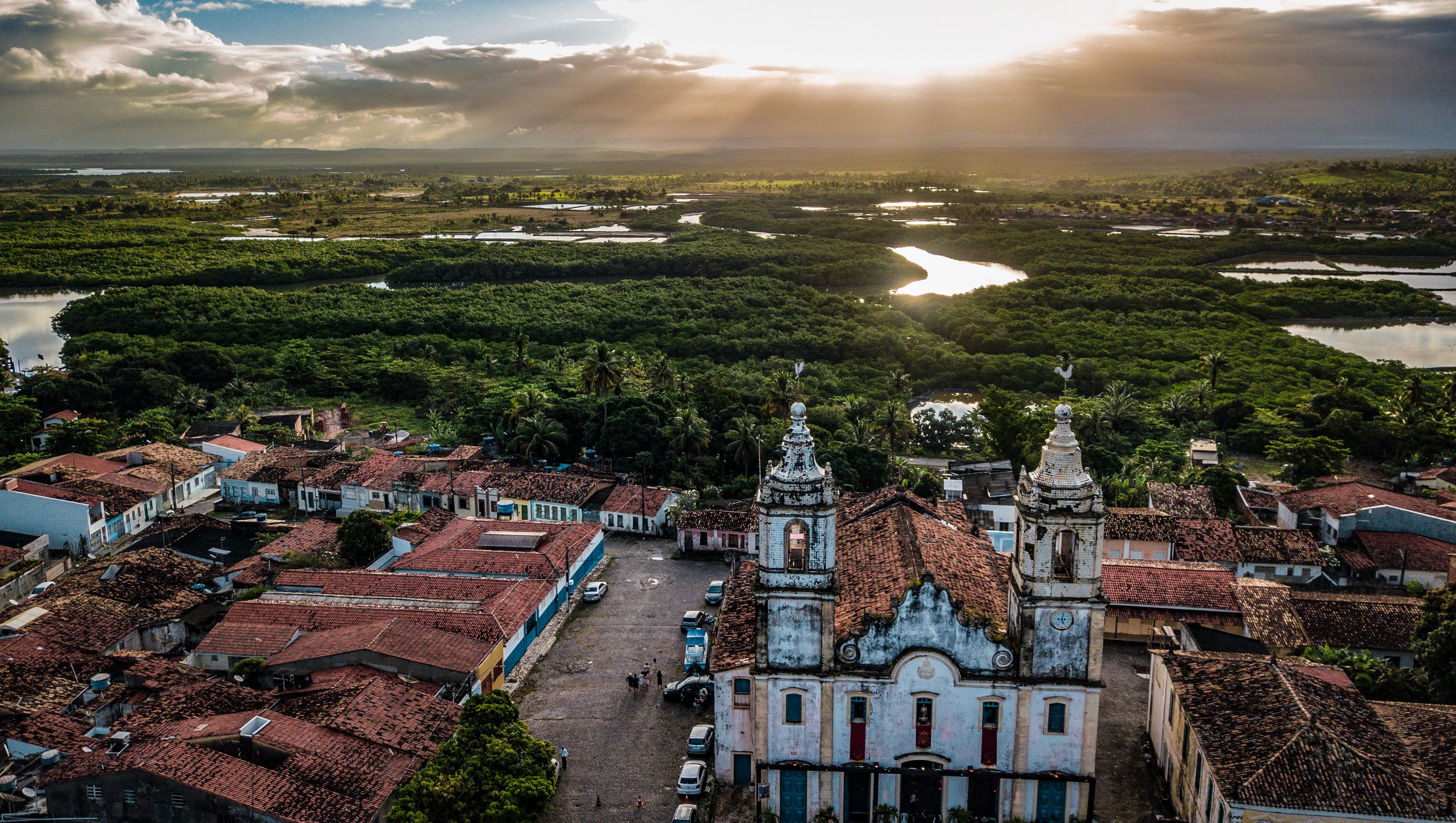 Vista aérea de São Cristóvão ao entardecer, com igreja de duas torres, telhados coloniais e manguezal ao fundo