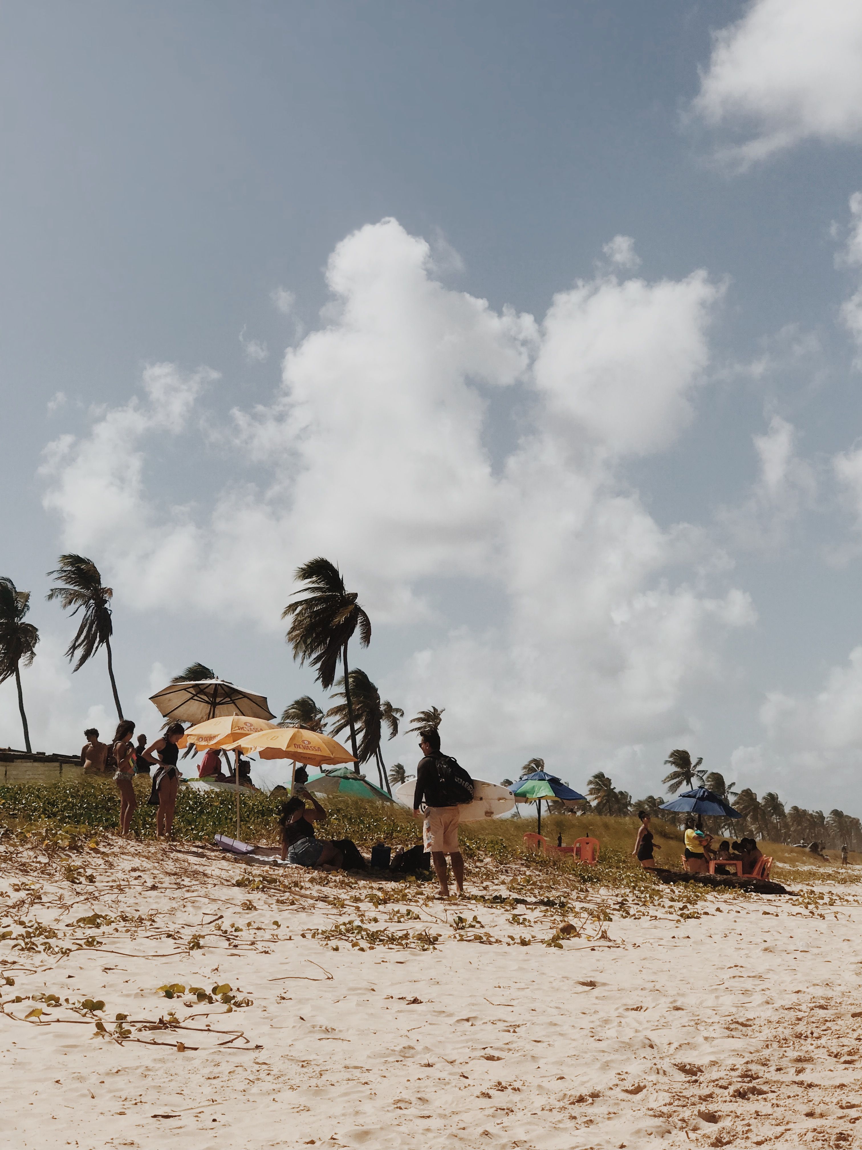 Praia de Atalaia em Aracaju, com coqueiros inclinados pelo vento, guarda-sóis coloridos na areia e pessoas na beira-mar