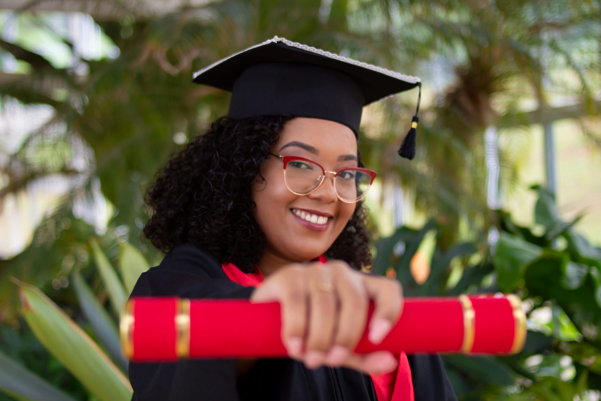 Formanda sorrindo com beca e canudo vermelho, num jardim.