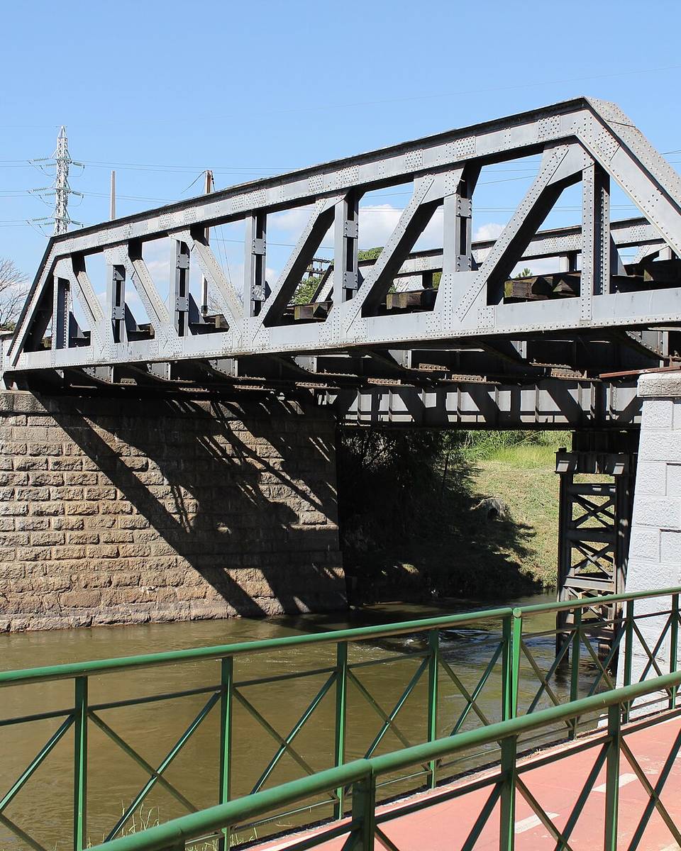 Ciclovia vermelha com grade verde à beira do rio Sorocaba, passando por baixo de uma ponte ferroviária de ferro.