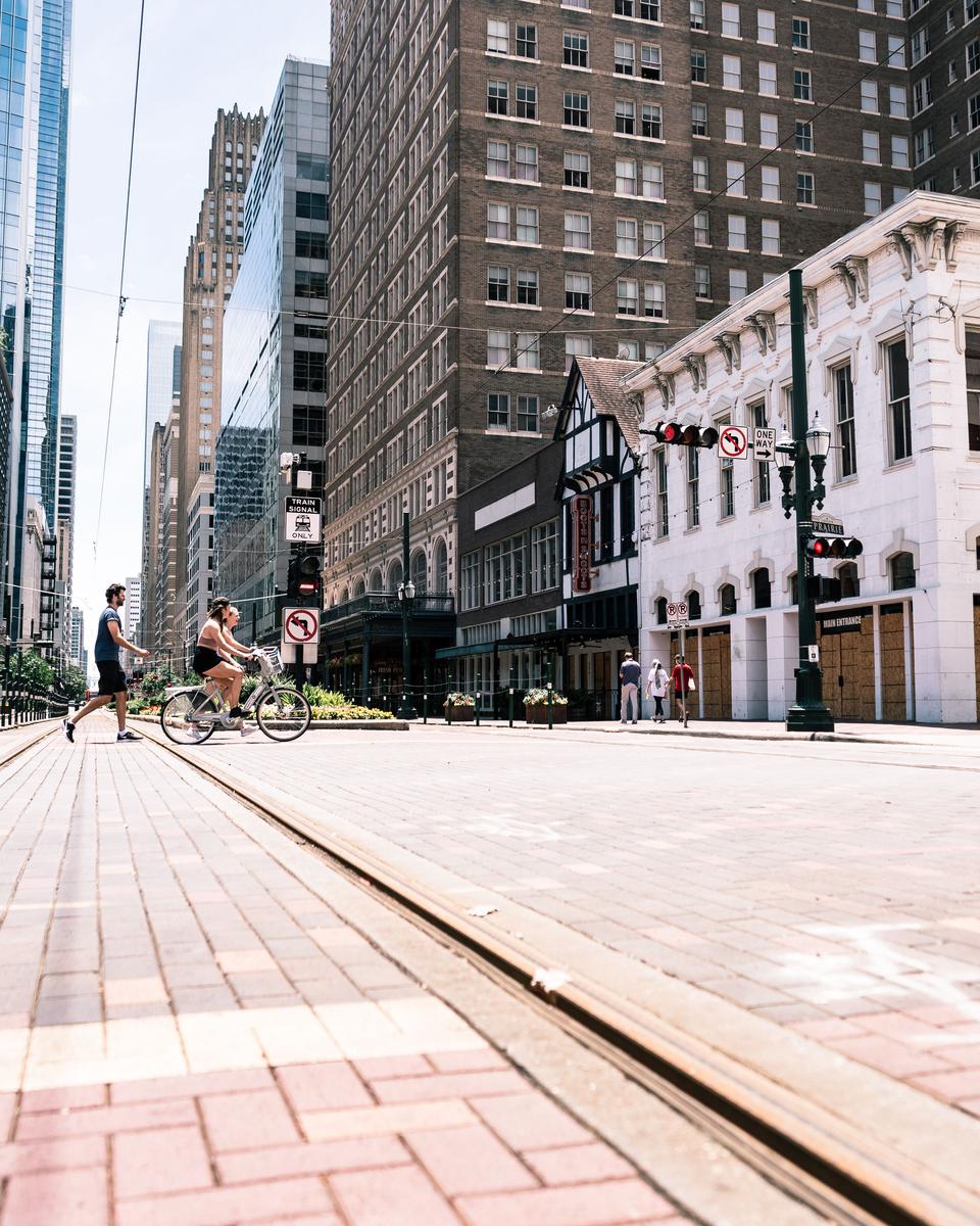 Rapaz atravessando a Main Street ao lado de uma ciclista, com os trilhos do metrô de superfície e os prédios antigos do centro de Houston.
