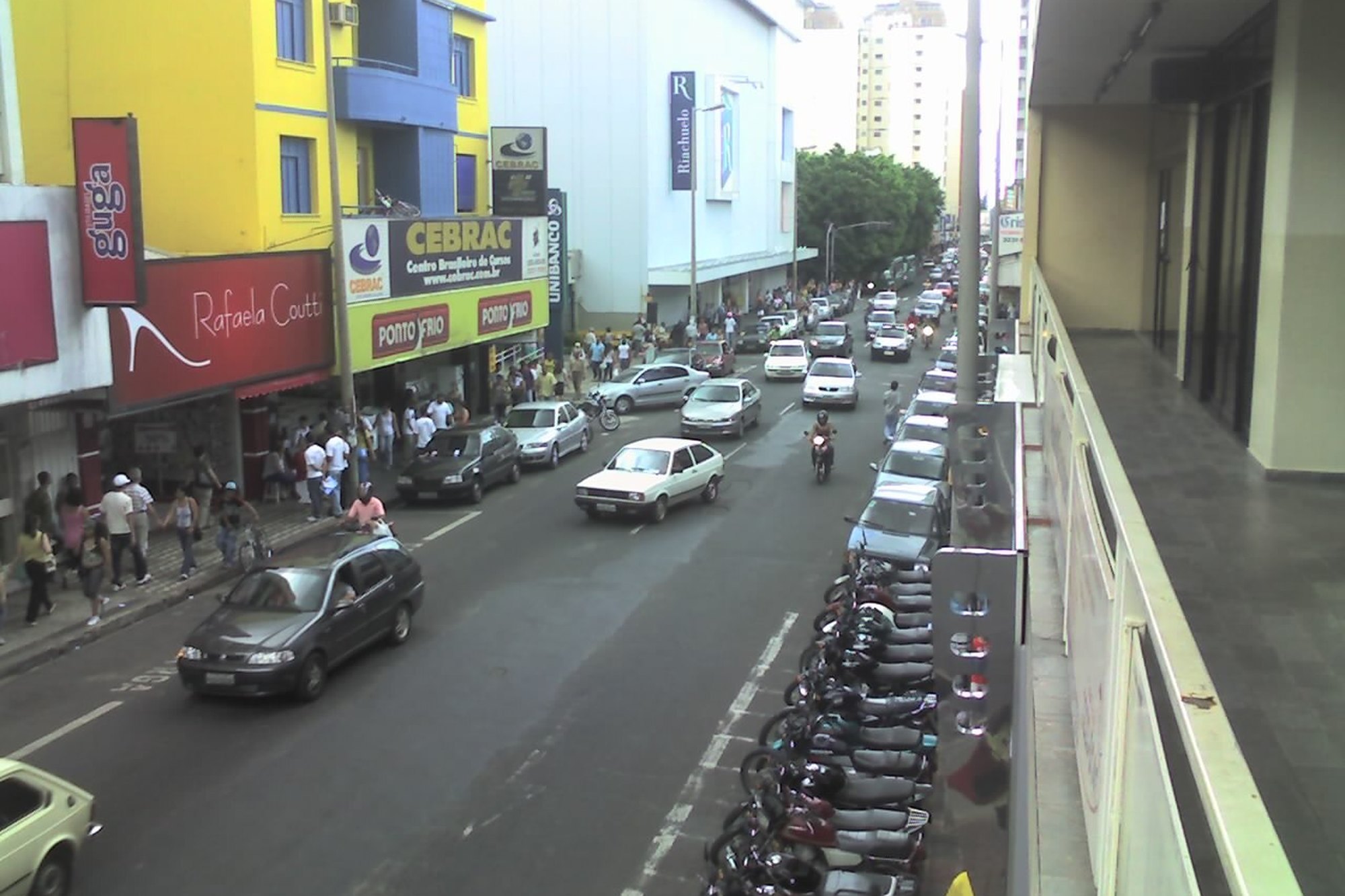 Rua comercial do Centro de Uberlândia vista do alto, com pedestres nas calçadas, fachadas coloridas de lojas, carros e uma fileira de motos estacionadas.